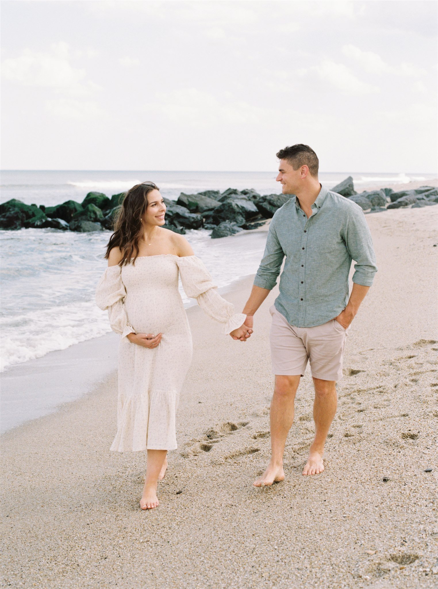 Pregnant woman cradling belly while holding hands with man on beach during Miami maternity session