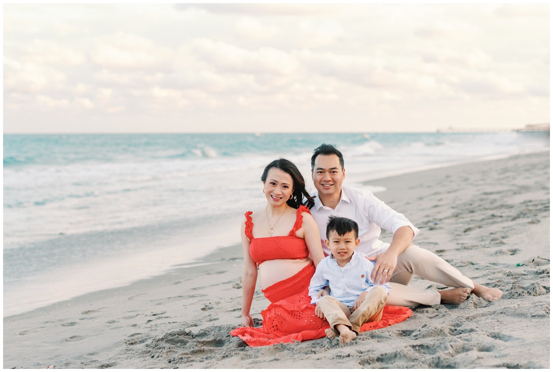 Woman wearing red top and skirt sitting next to husband and son on the beach during maternity photos
