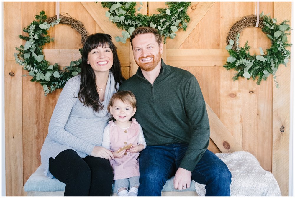 Mom, dad, and toddler sitting on bench in front of holiday wreaths