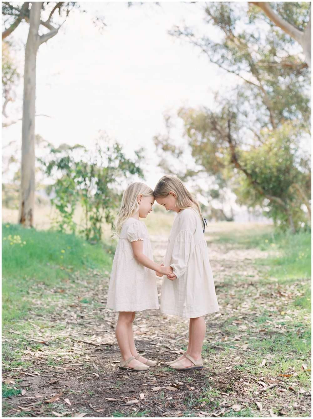 Sisters wearing white dresses, holding hands, and touching heads during session with NKB Photo