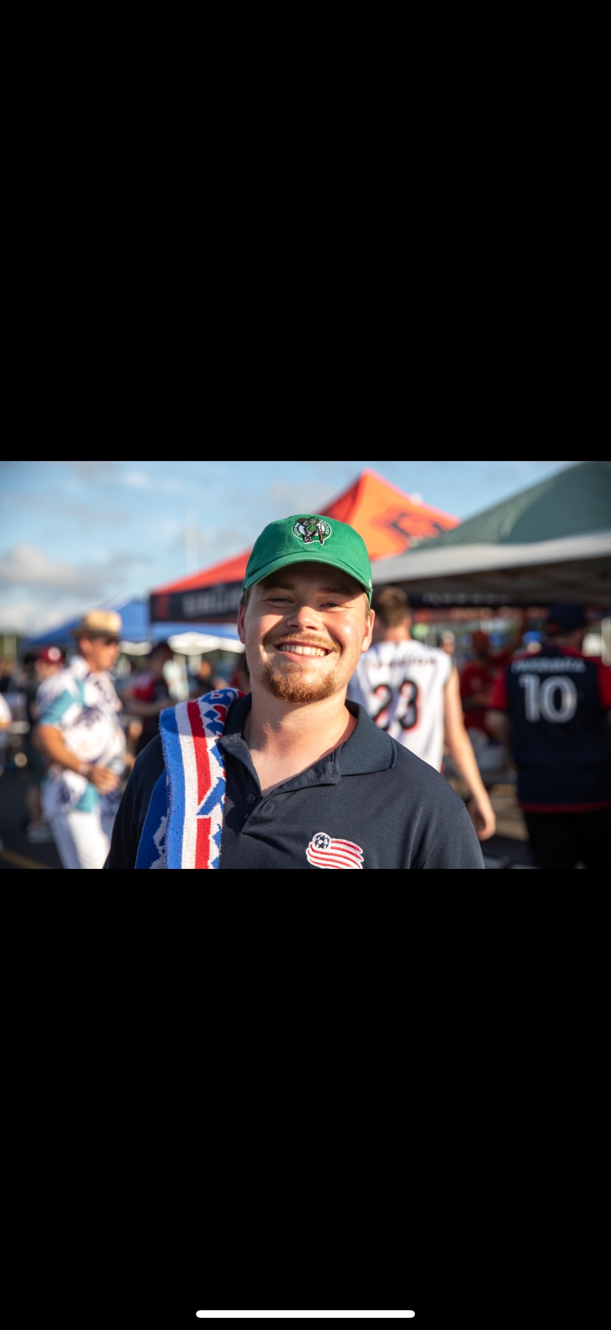 A young man smiling at a sports event, wearing a green baseball cap with a logo, a gray polo shirt with a travel sticker, and holding a towel with red, white, and blue stripes. There are people and tents in the background, indicating an outdoor gathering.