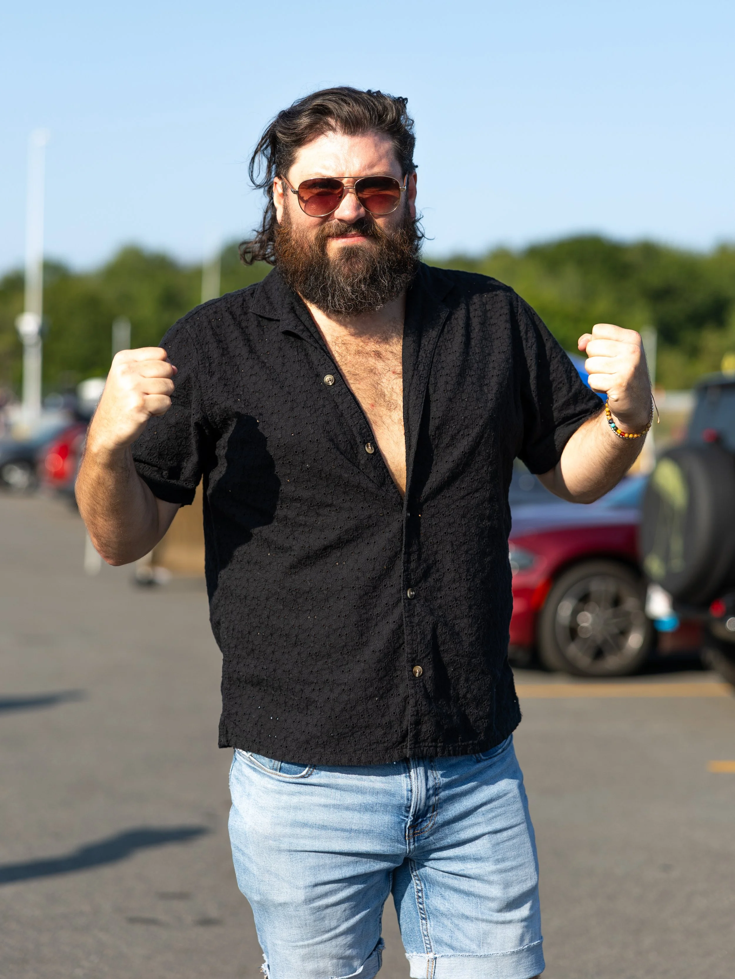 A man with long dark hair, a beard, and sunglasses flexes his biceps outdoors on a sunny day, wearing a black short-sleeve shirt and light blue shorts.