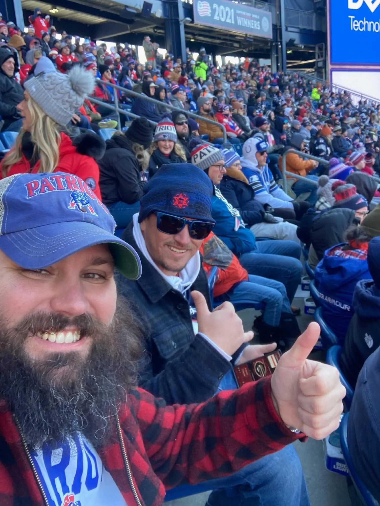 Crowd of sports fans seated in stadium stands, wearing hats and scarves, with two men in the foreground showing thumbs up and smiling for the camera, one wearing sunglasses and a beanie, the other with a baseball cap.
