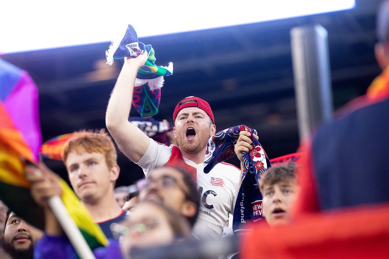 A group of sports fans cheering, with one man in a white jersey and red cap raising a scarf, shouting enthusiastically.