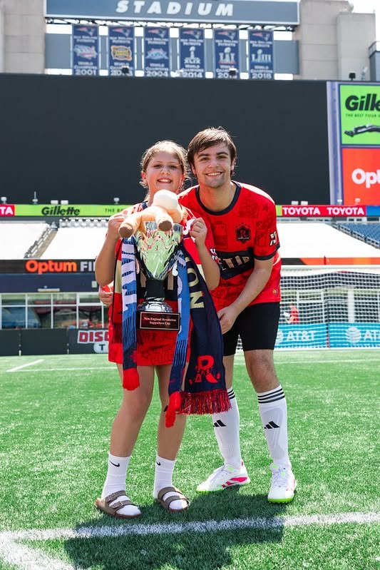 A young girl and a man are standing together on a soccer field inside a stadium, smiling at the camera. The girl is holding a trophy and a small stuffed animal, wearing a red soccer uniform and sandals. The man is wearing a red and black soccer jersey and white socks with black stripes, leaning slightly toward the girl.