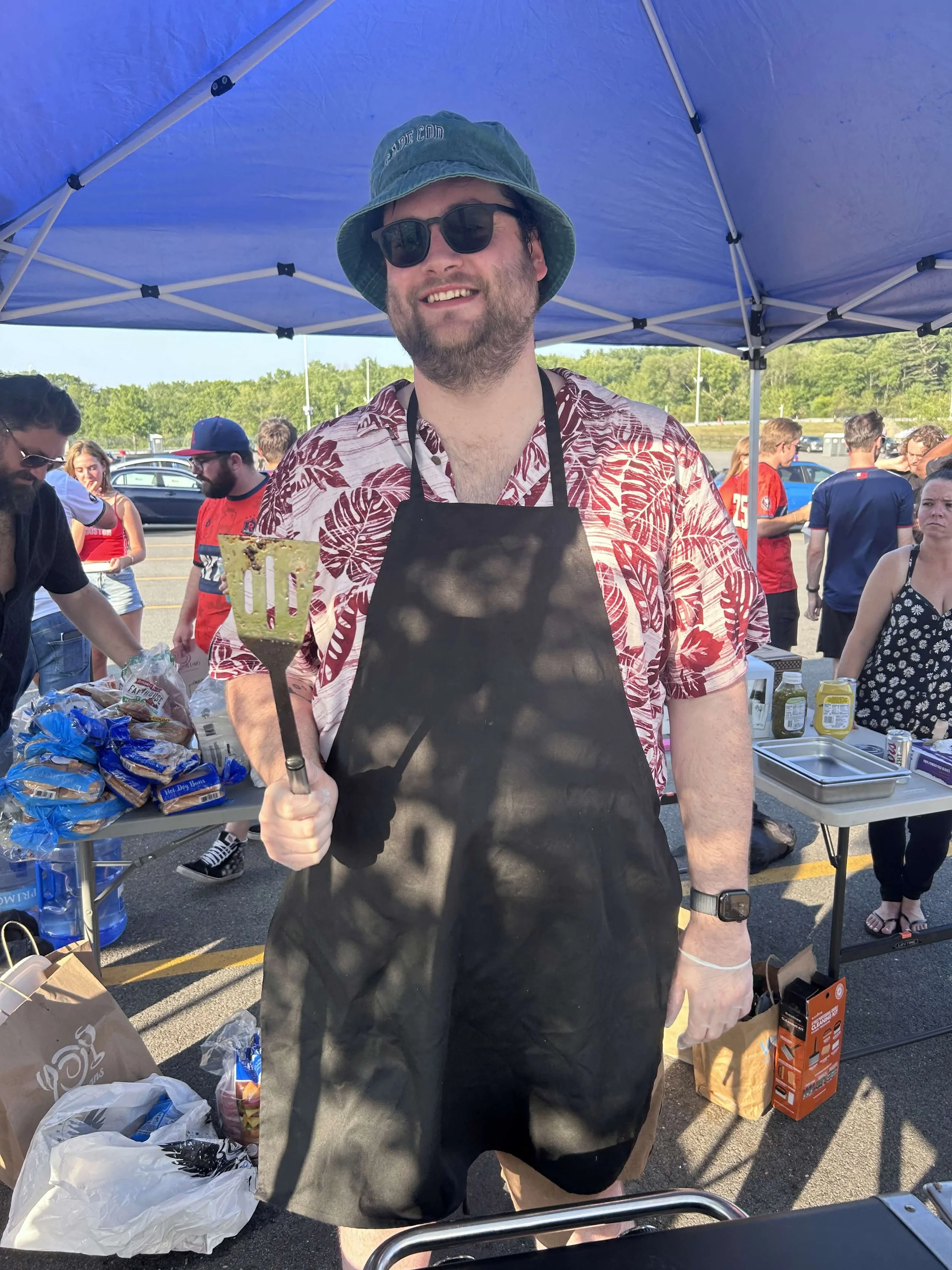 A man wearing sunglasses, a bucket hat, a black apron, and a Hawaiian shirt is smiling while holding a spatula at an outdoor event under a blue canopy, with several people and tables of food in the background.