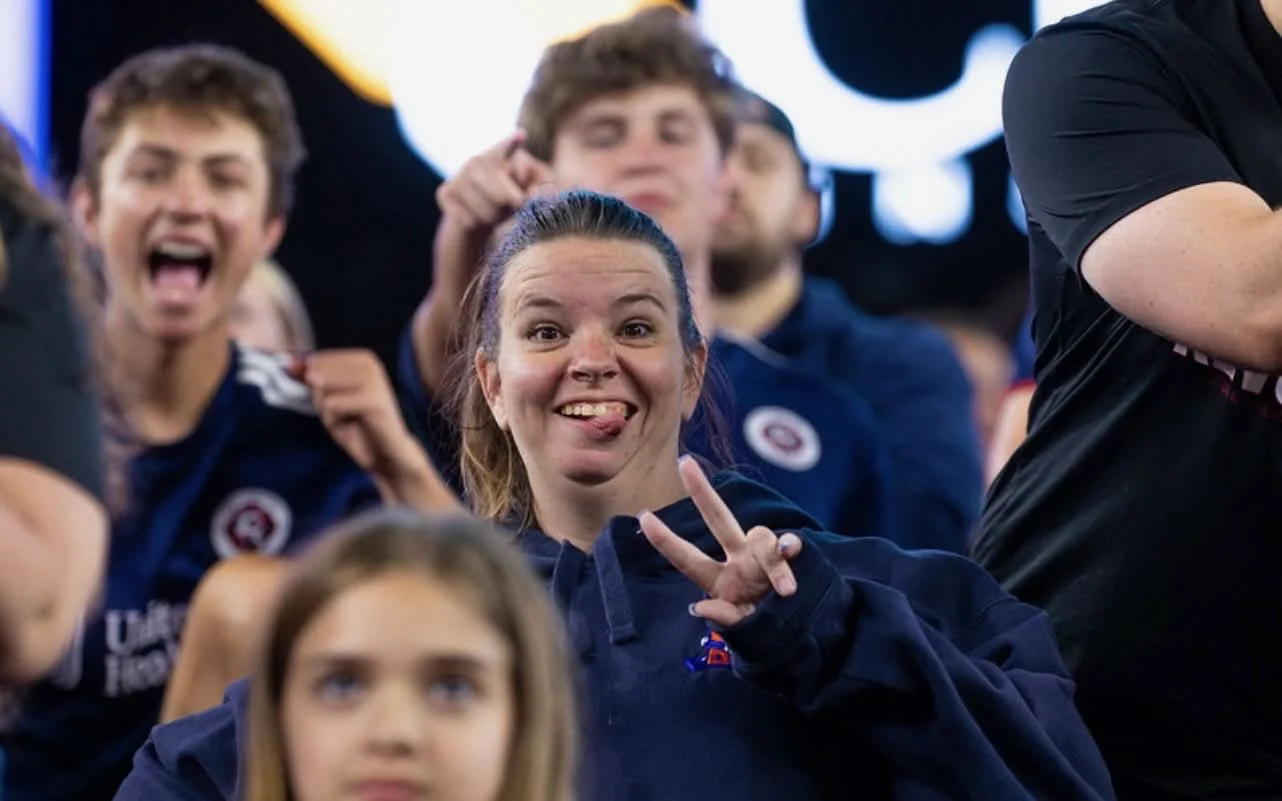 Group of young people at a sports event, with a woman in the center making a peace sign and sticking out her tongue, smiling and playful, wearing dark clothing, others smiling and cheering in background.