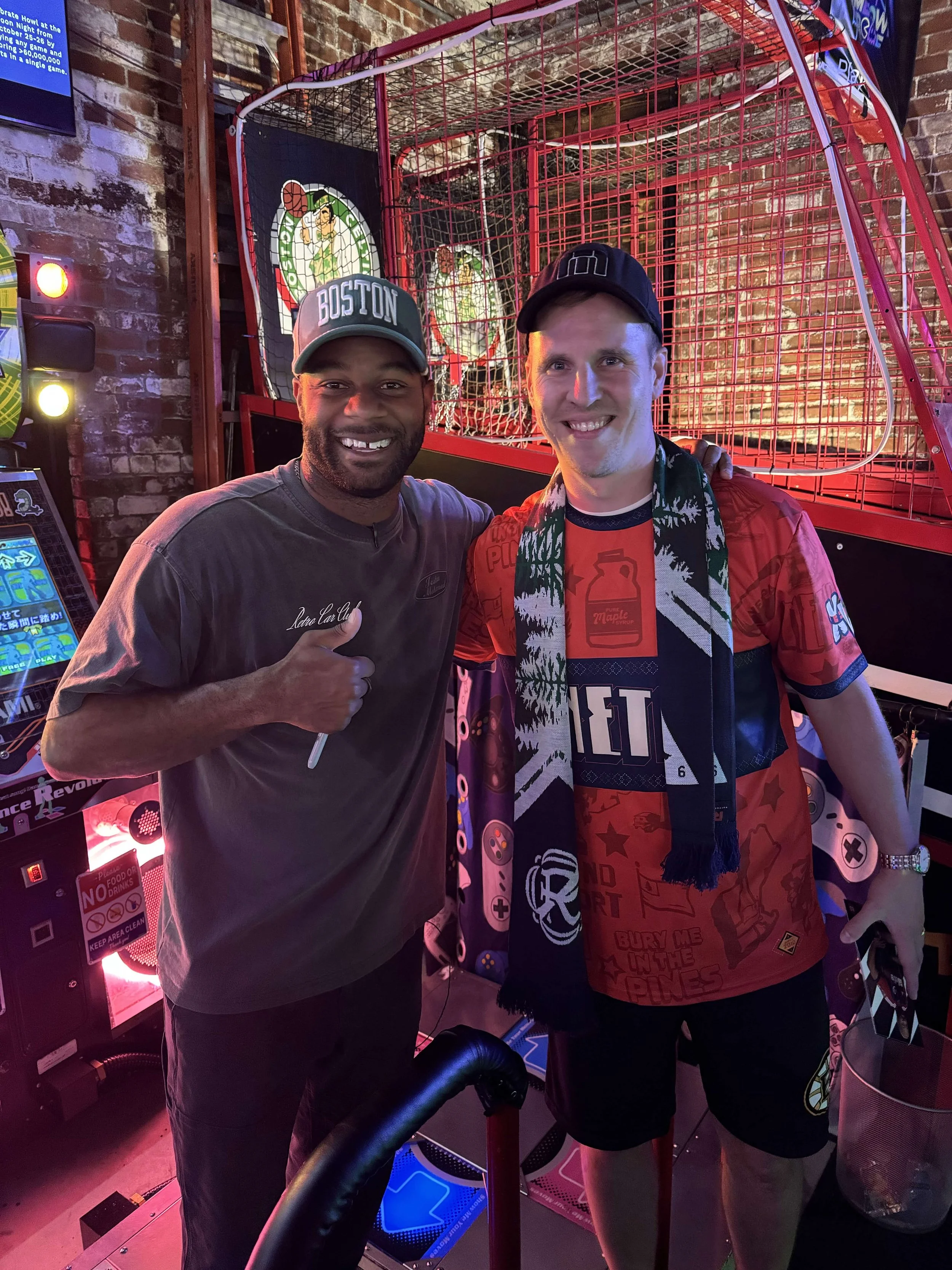 Two smiling men standing together in an arcade, one pointing at the camera, with arcade machine and sports-themed decor in the background.