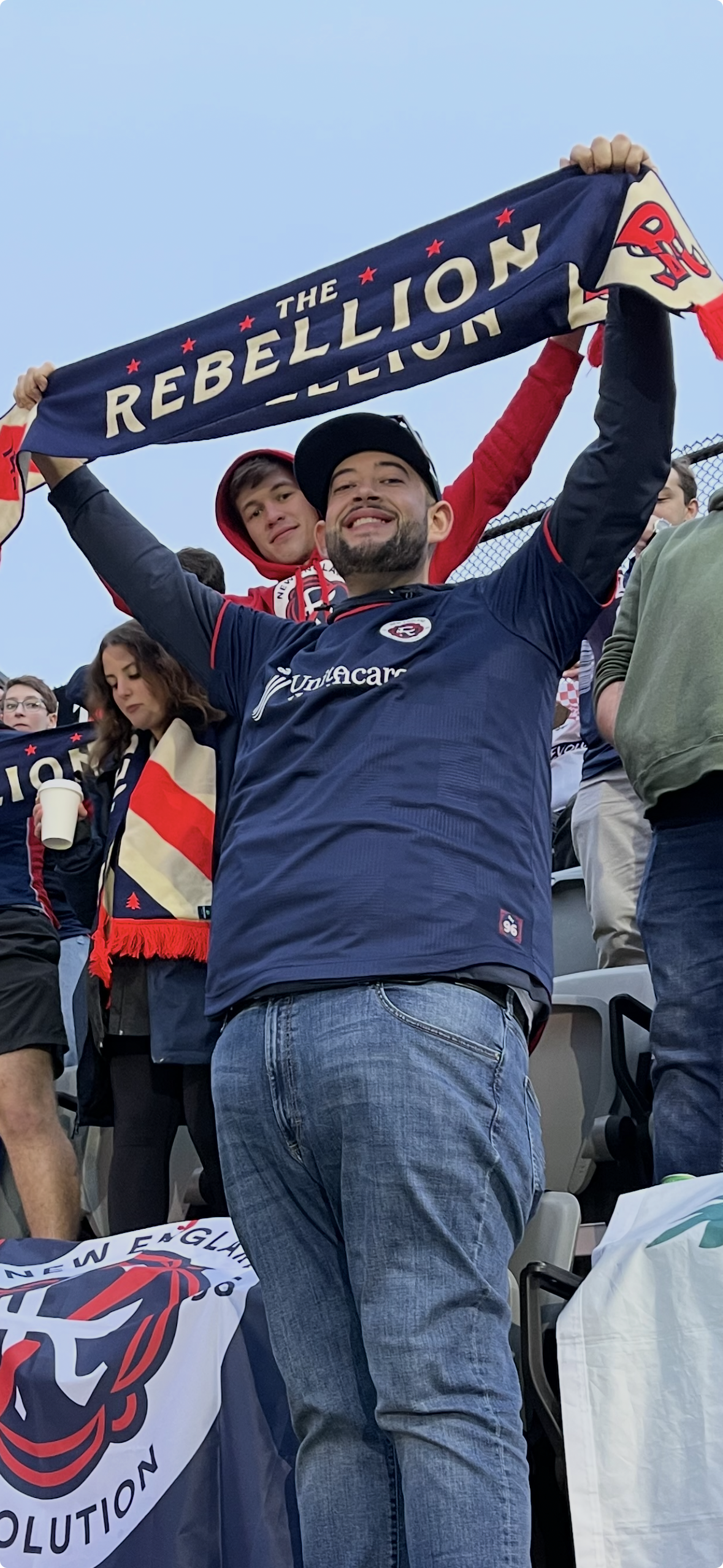 A group of sports fans at a stadium, with one man in the foreground holding a scarf that says "The Rebellion" and smiling. Other fans around him are wearing team apparel and holding beverages.