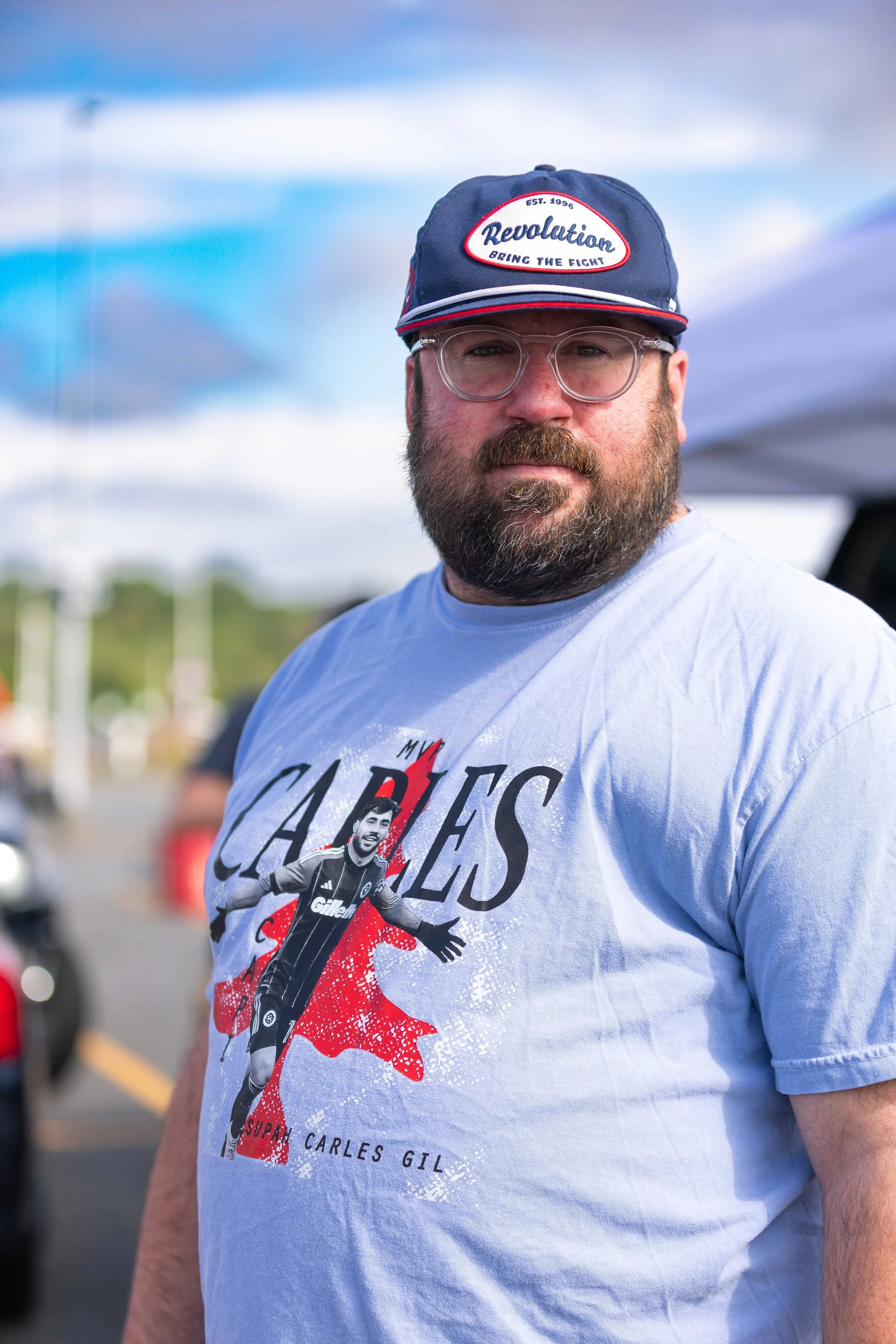 A man with glasses and a beard wearing a navy blue cap and a light blue t-shirt with a graphic of a soccer player and the words 'Cafes Gil' at an outdoor event.