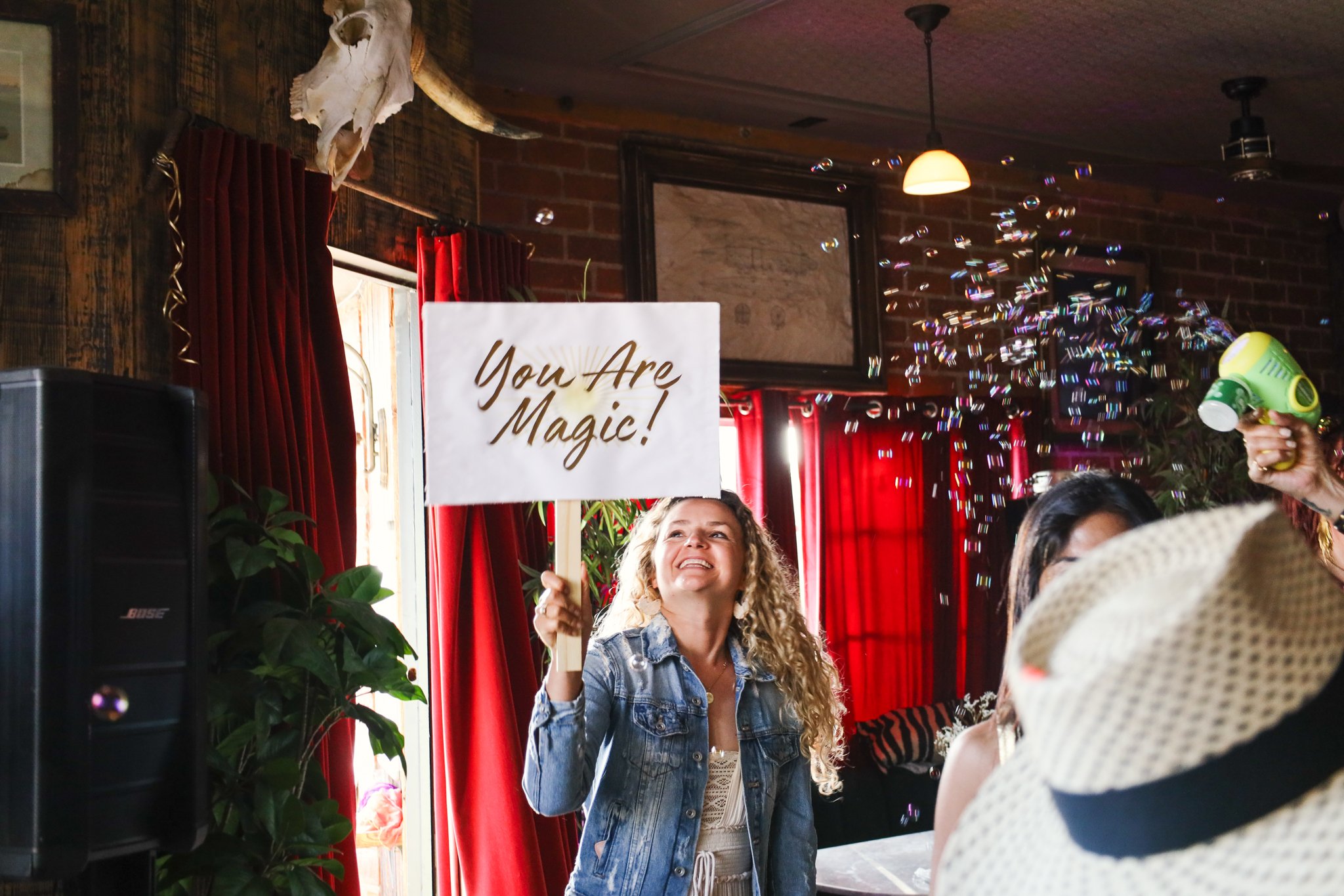Woman smiling and holding a sign that says "You Are Magic!" at a celebration or party with bubbles and party decorations in the background.