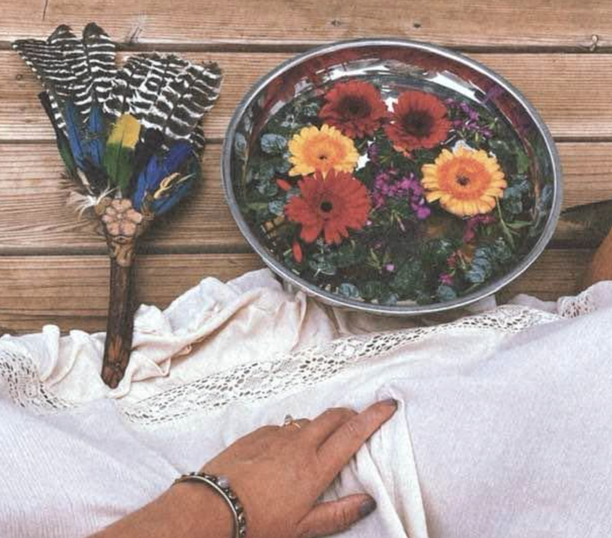 A hand resting on a white lace-trimmed fabric with a silver bracelet, a metal bowl filled with colorful floating flowers, and a butterfly-shaped decorative fan with vibrant blue, black and green feathers on a wooden surface.