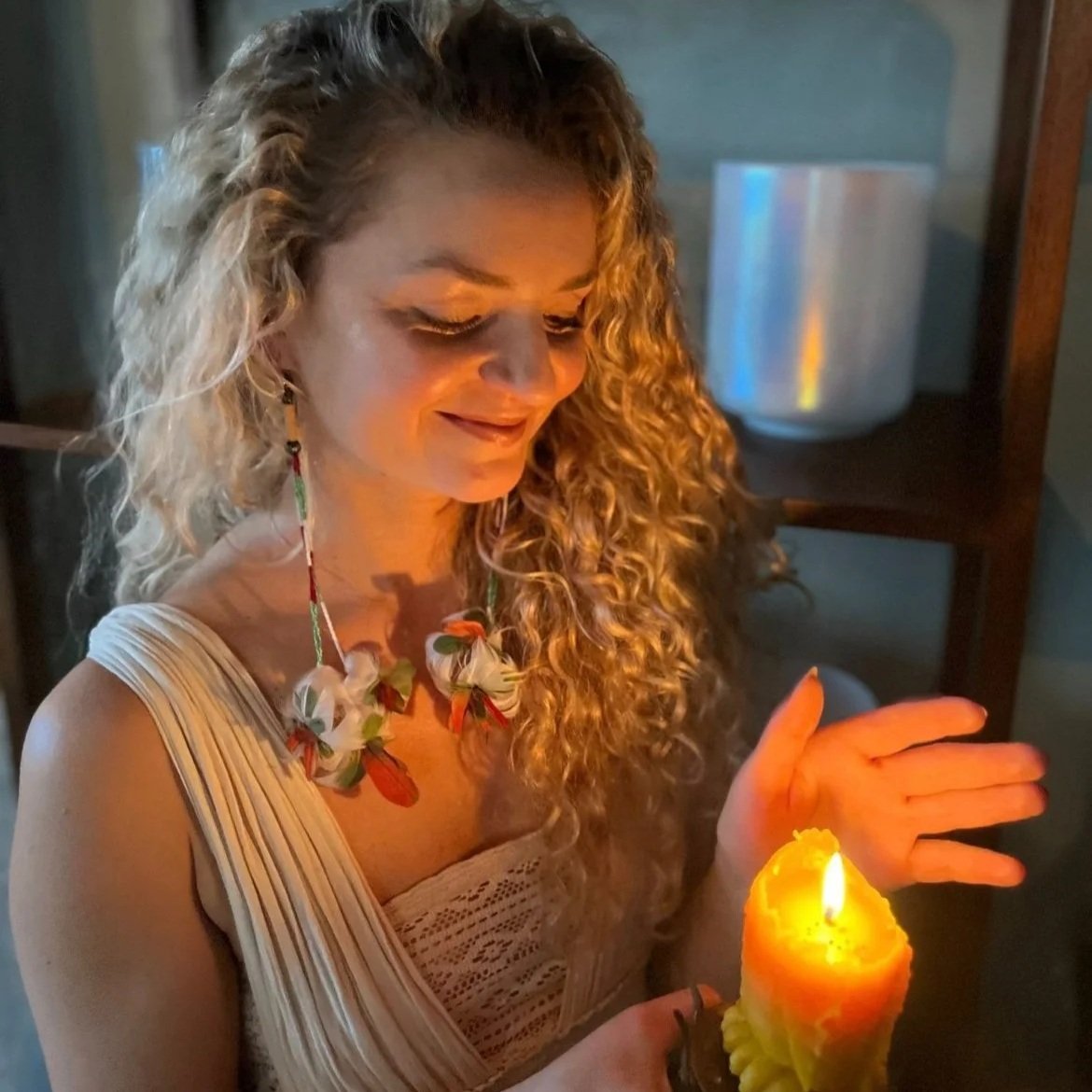Woman with curly blonde hair, wearing a floral necklace and a beige dress, holding a lit candle in a decorative holder, smiling softly, in dim lighting.