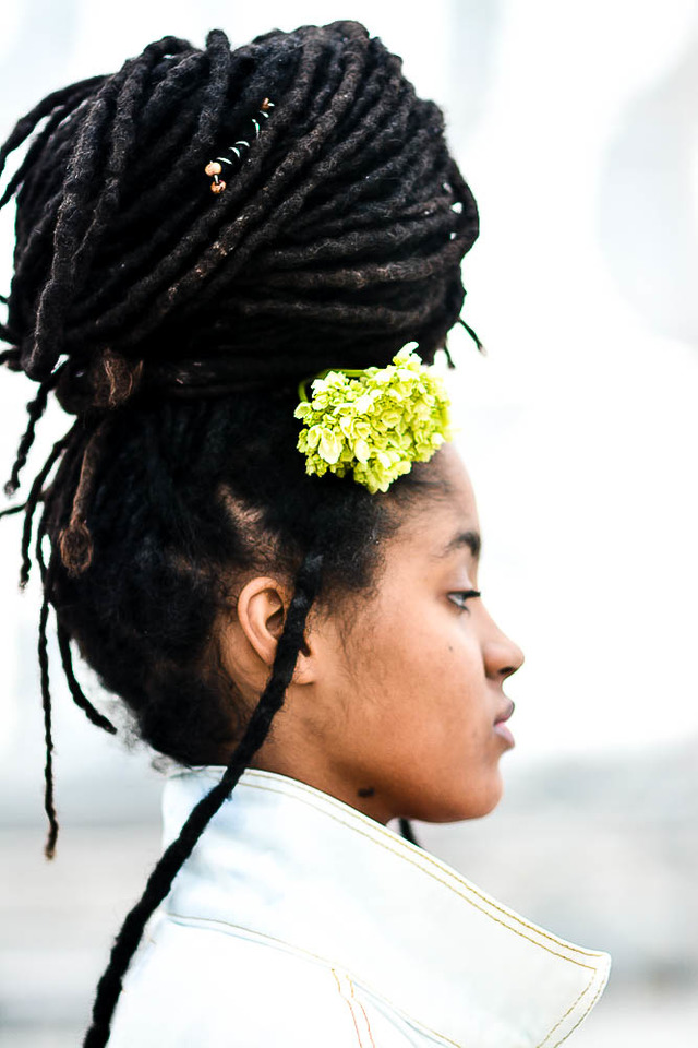 A woman with a flower in her dreadlocked hair looks of into the distance. 