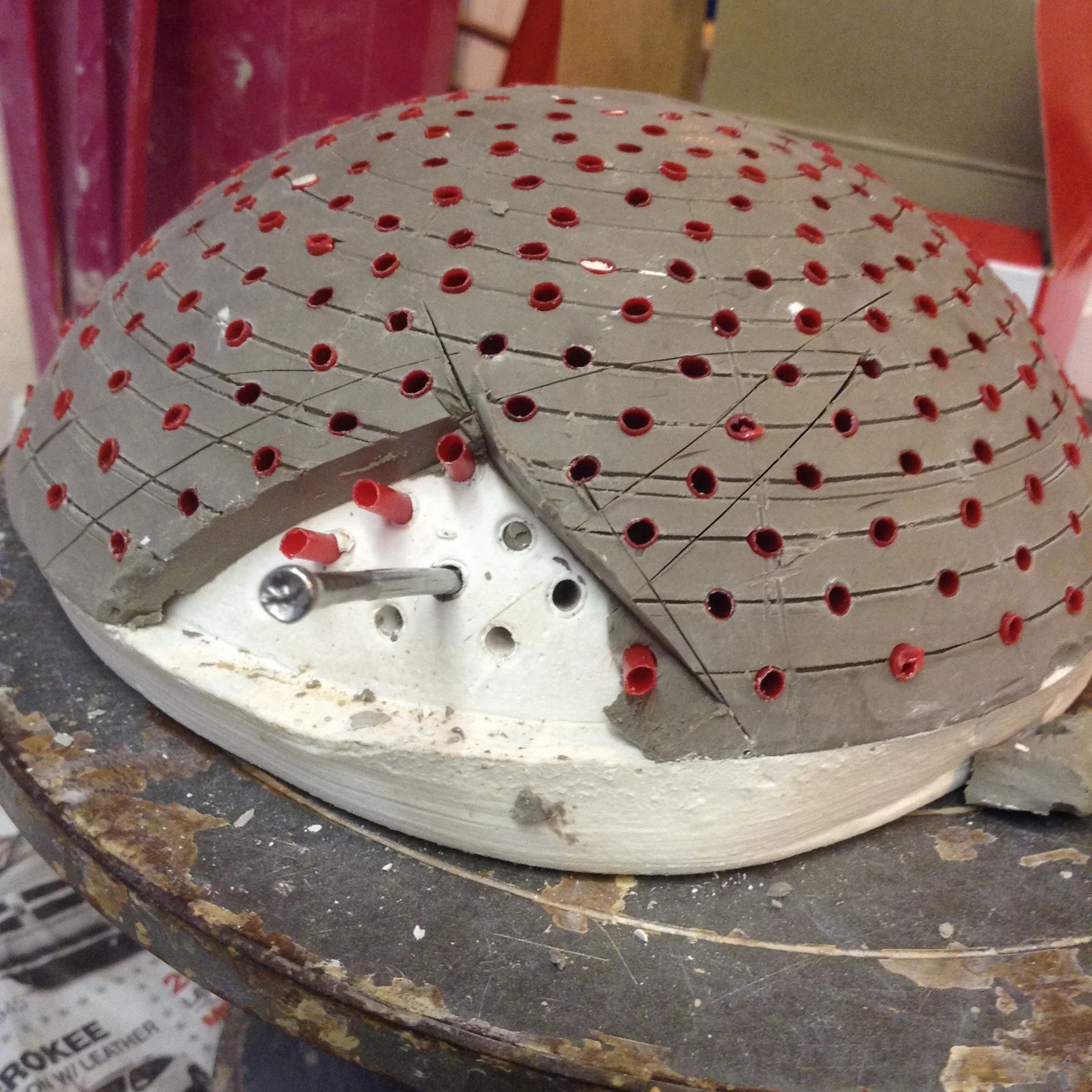 Unmolding process of plaster dome form. Clay form was sculpted for pouring plaster inside. Lines were drawn on outside of clay bowl for locations of red coffee stirrers to push through. These red pieces are encased in plaster, but pull out, solving the problem of my not having a drill press. Here, the wet clay bowl form is being dismantled after the plaster has set. The metal nail is shown here as example of the radiating angle all the holes have.