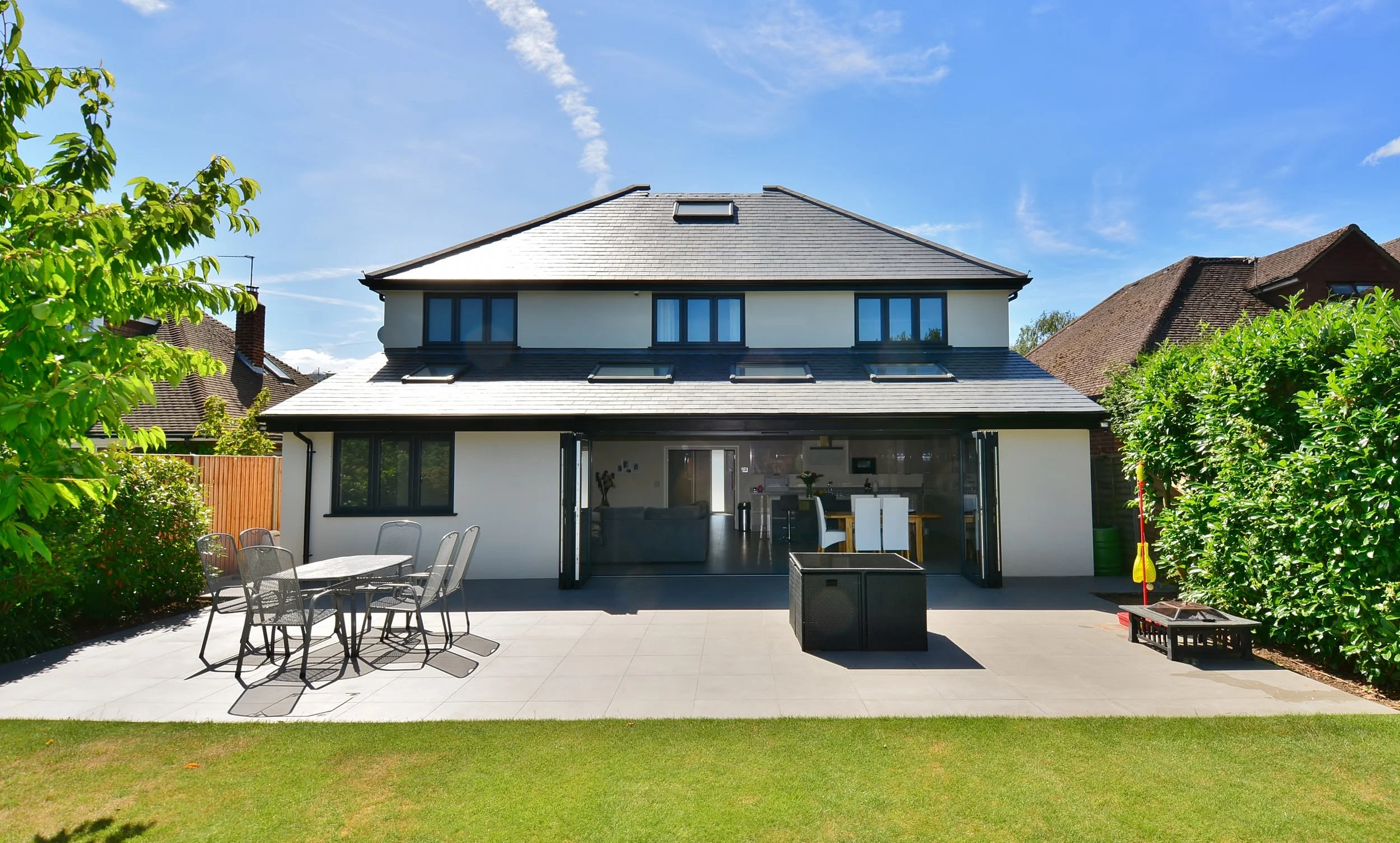 Modern two-story house with open patio and outdoor furniture, surrounded by greenery on a sunny day.