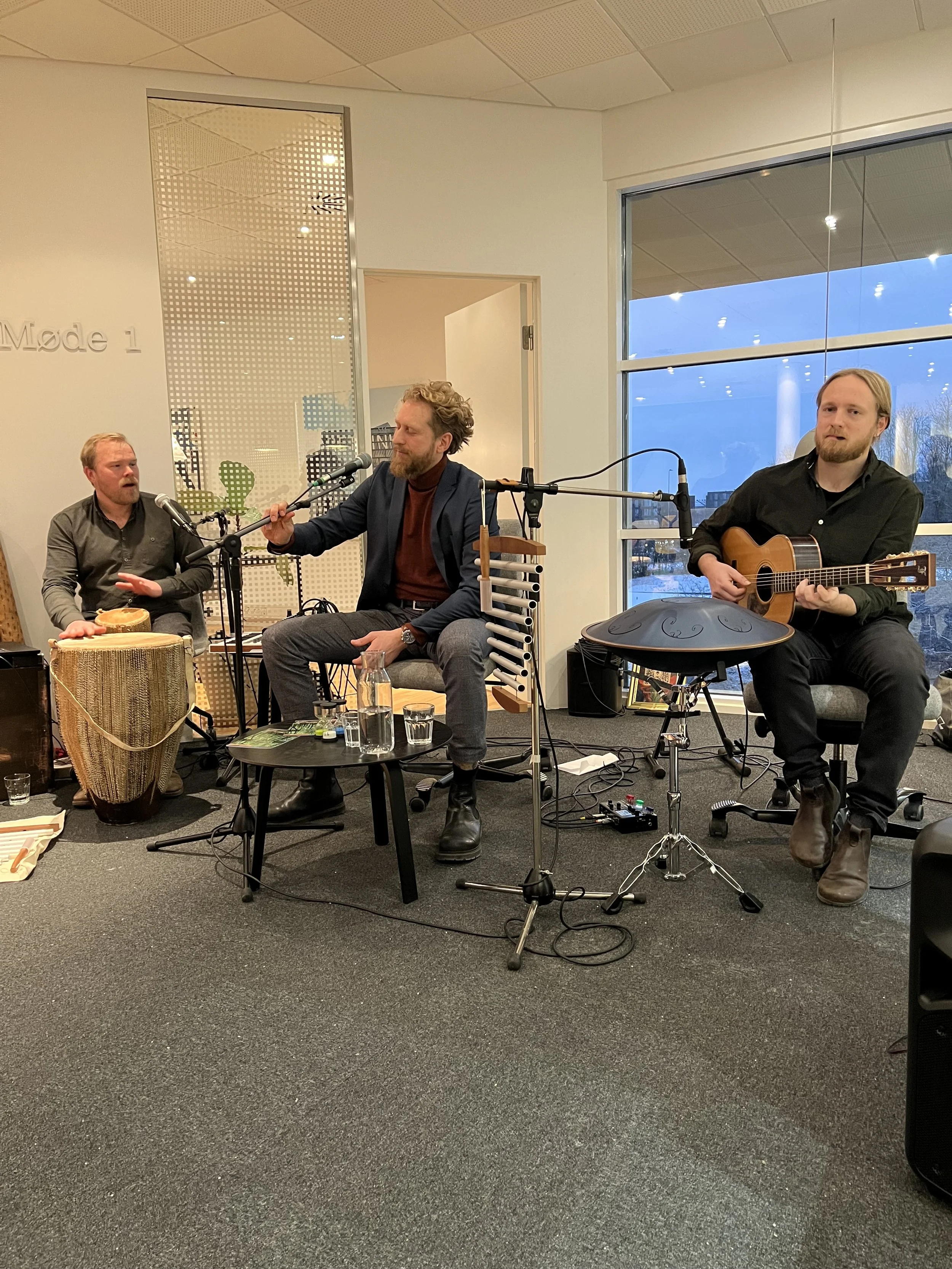 Tre musikere spiller akustisk musik i et lyst rum med store vinduer. Der er mikrofoner, instrumenter, og små borde med drikkevarer foran dem.