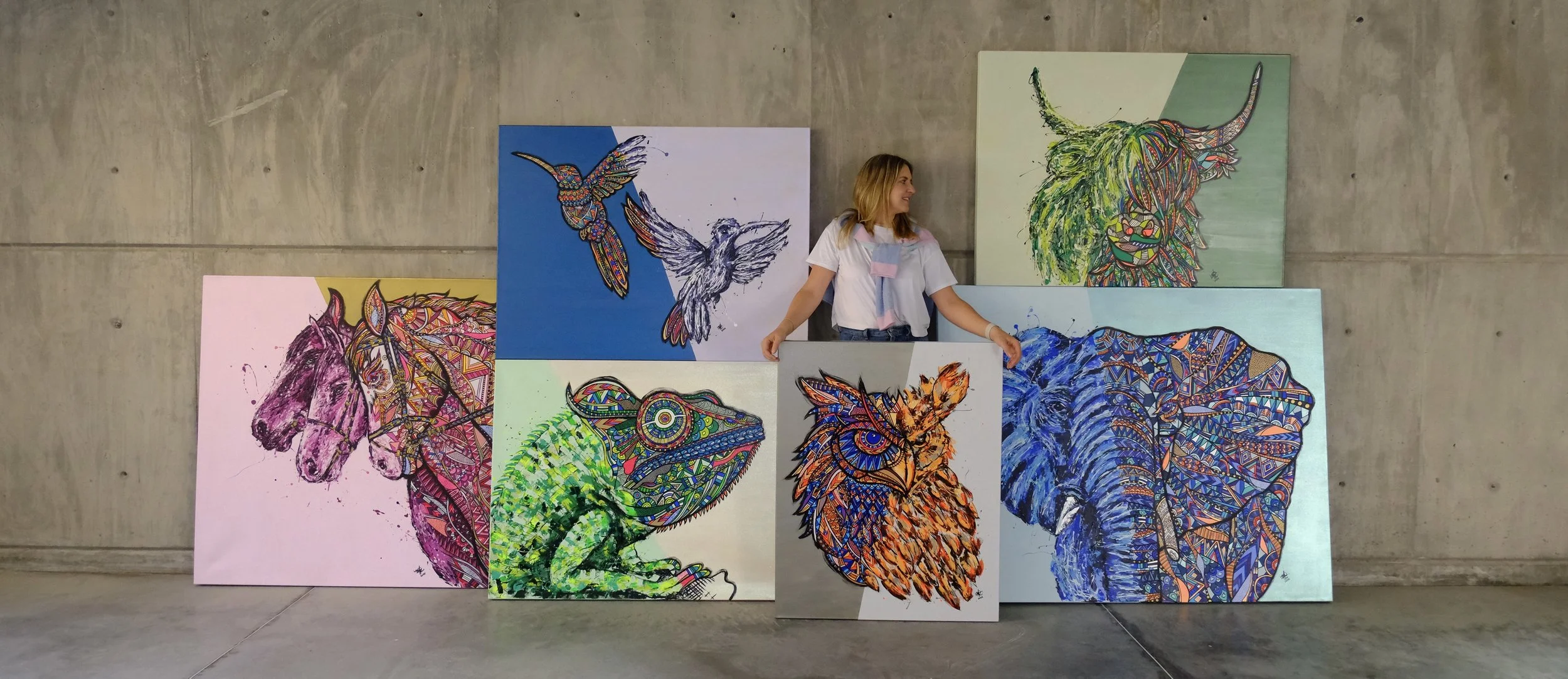 Artist standing in front of seven colorful animal portraits on canvas leaning against a concrete wall, including a horse, owl, chameleon, lion, elephant, and a two hummingbirds.