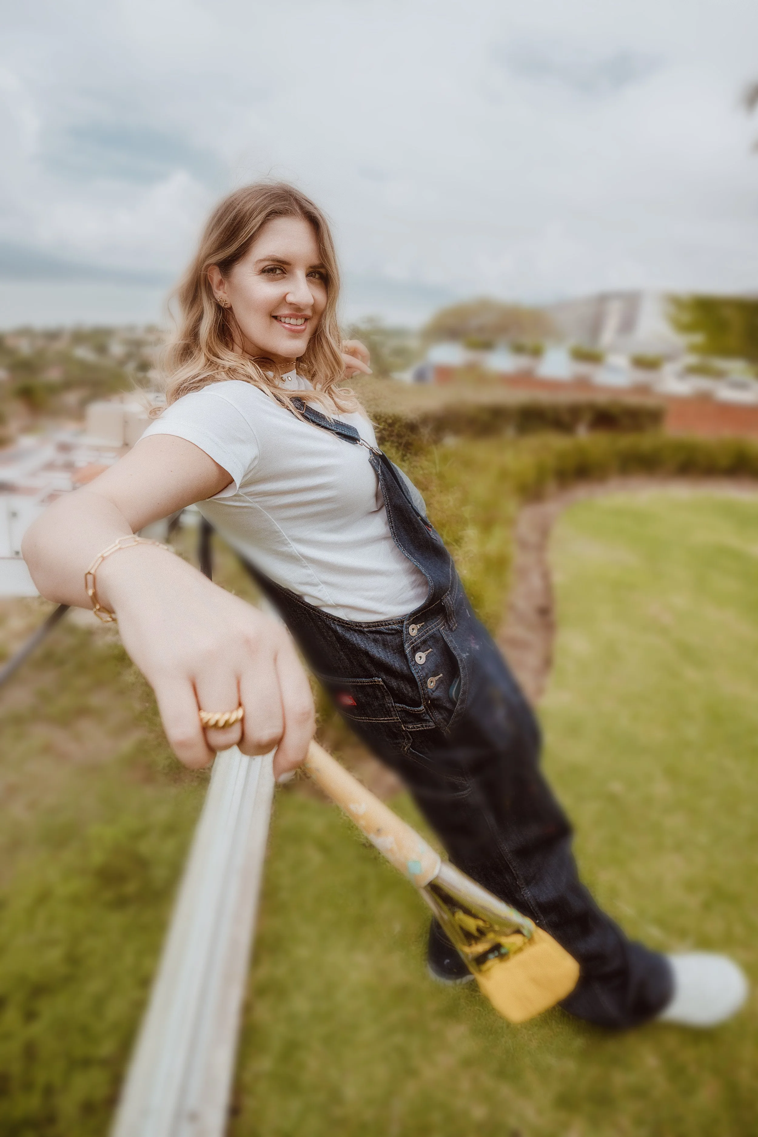 Young woman smiling while holding a selfie stick on a rooftop garden with a cityscape and cloudy sky in background.