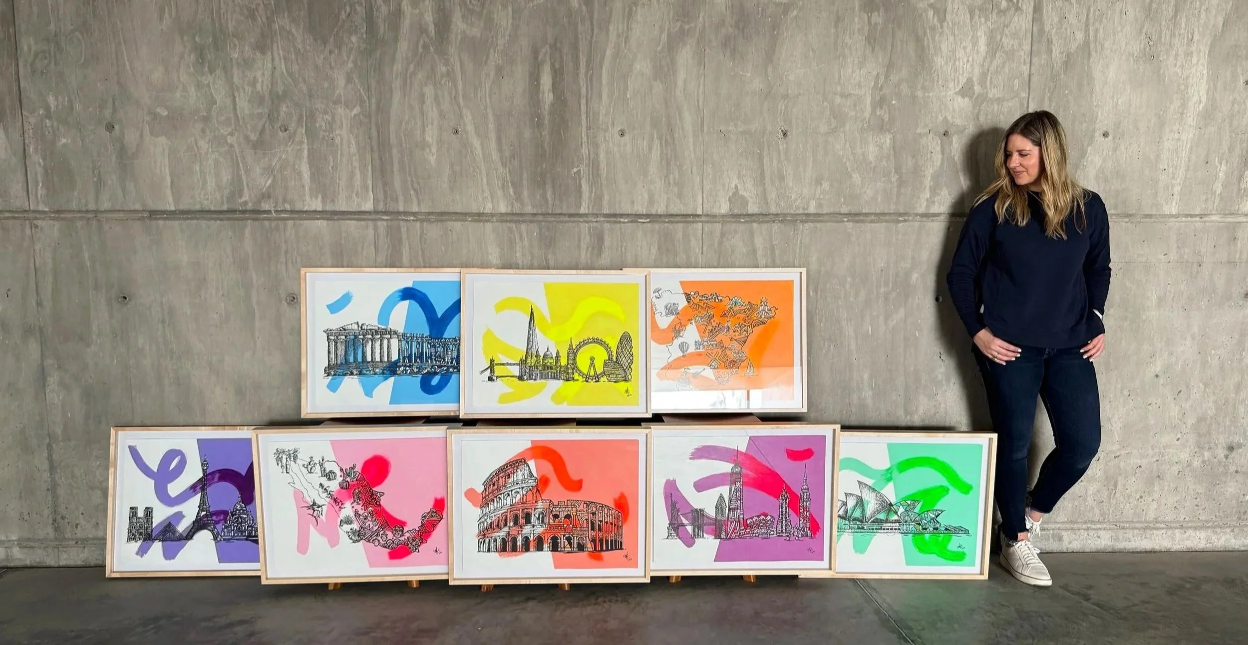 A woman with blond hair, dressed in a dark blue sweatshirt and black jeans, leaning against a concrete wall, stands next to eight framed artworks on the ground. The artworks feature black line drawings of famous landmarks overlaid with colorful brush strokes in various colors like blue, yellow, orange, pink, purple, and green.