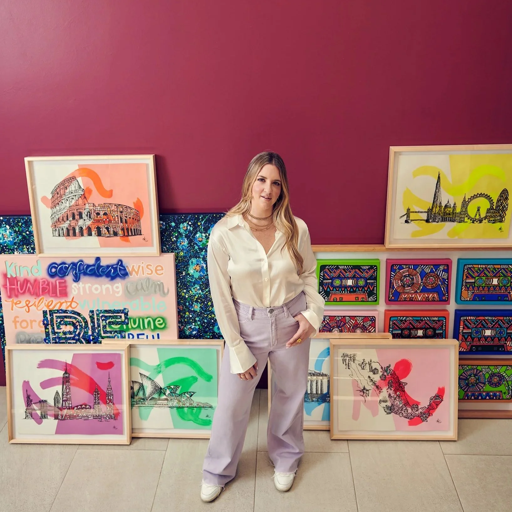 Woman in white blouse and light pants standing in front of colorful artwork depicting famous landmarks and cityscapes.