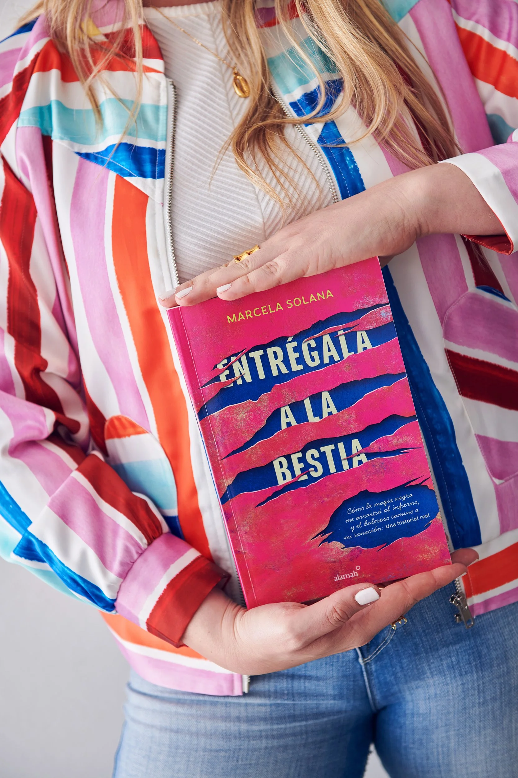 A woman holding a bright pink book titled 'Entrégala a la Bestia' with torn paper design on the cover. She is wearing a multicolored striped jacket and a white top, with blonde hair, a gold necklace, and a ring visible on her hand.
