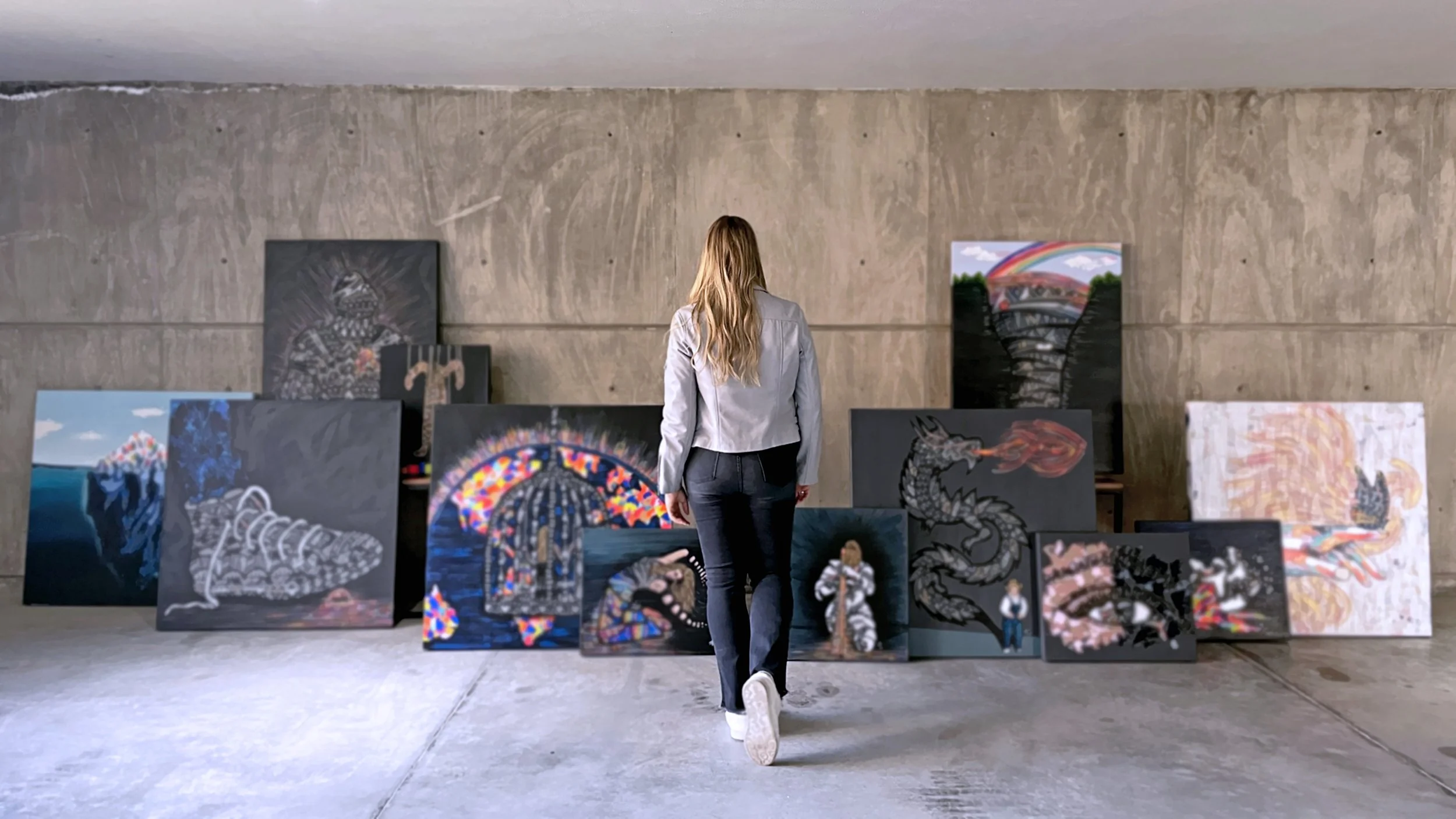 A woman is walking toward a collection of colorful paintings arranged against a concrete wall in an art gallery or studio.