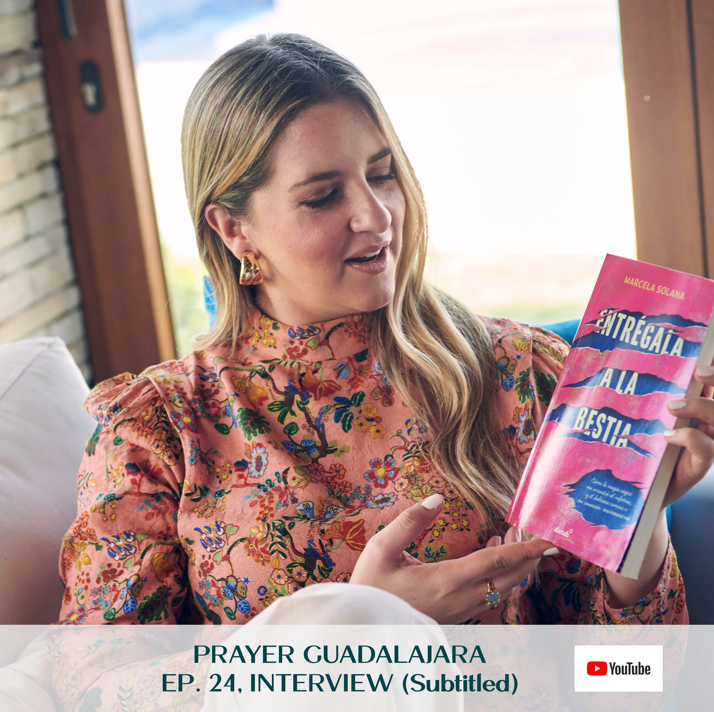 Marcela Solana smiling and holding her pink book titled 'Entregala a la Bestia' by Marcela Solana. She is indoors with a background of a window and light streaming in, wearing a floral blouse and gold earrings.