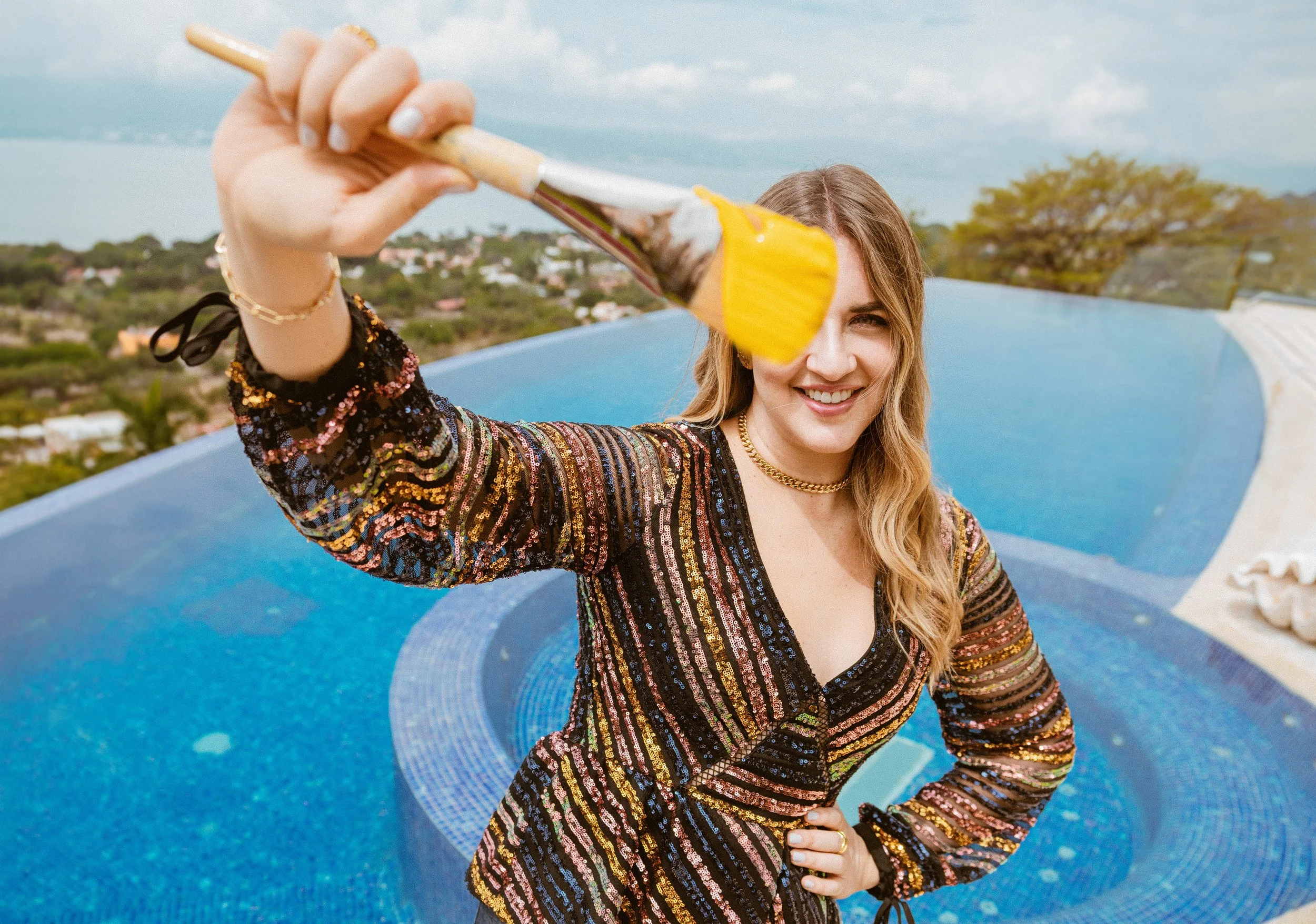 A woman wearing a black, long-sleeved, sequined dress with multicolored stripes, smiling and holding a paintbrush with yellow paint close to the camera. She is standing by a blue swimming pool with a scenic landscape and cloudy sky in the background.
