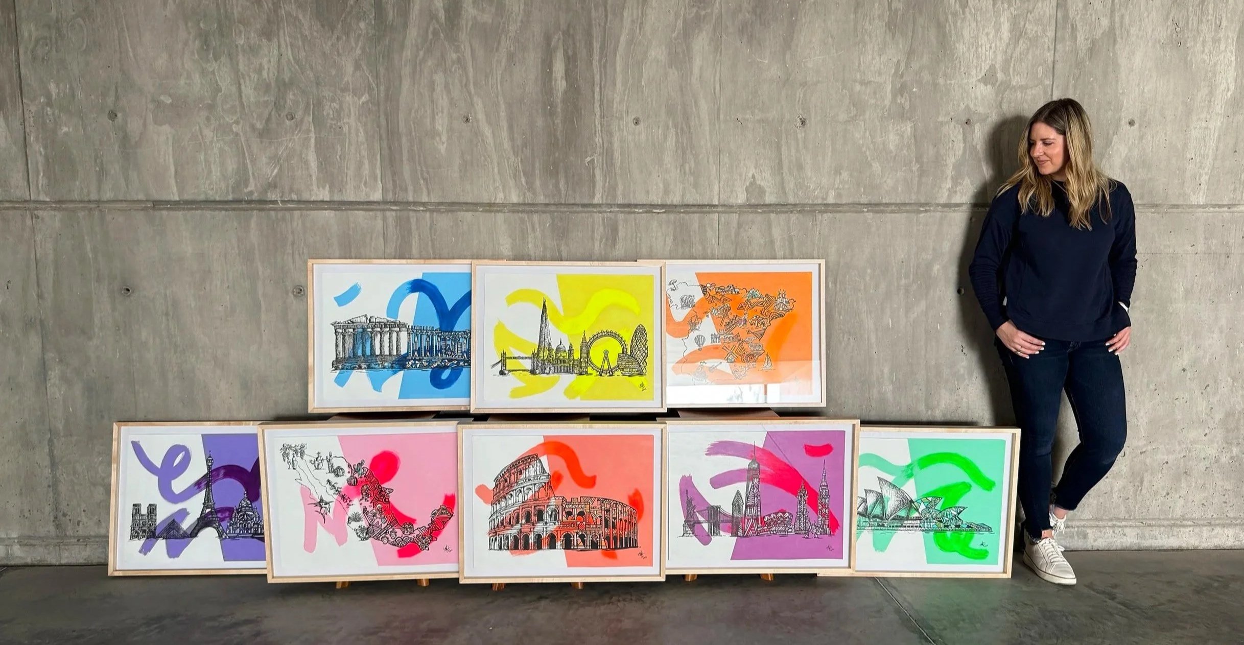 A woman standing next to eight framed art prints displayed against a concrete wall. The prints feature line drawings of famous landmarks with colorful abstract backgrounds, including the Parthenon, Big Ben, the Colosseum, the Sydney Opera House, and others.