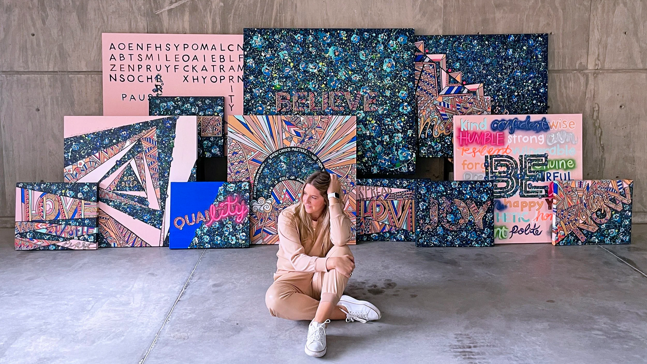 A woman sitting cross-legged on the floor, surrounded by colorful abstract and motivational artwork displayed against a concrete wall.