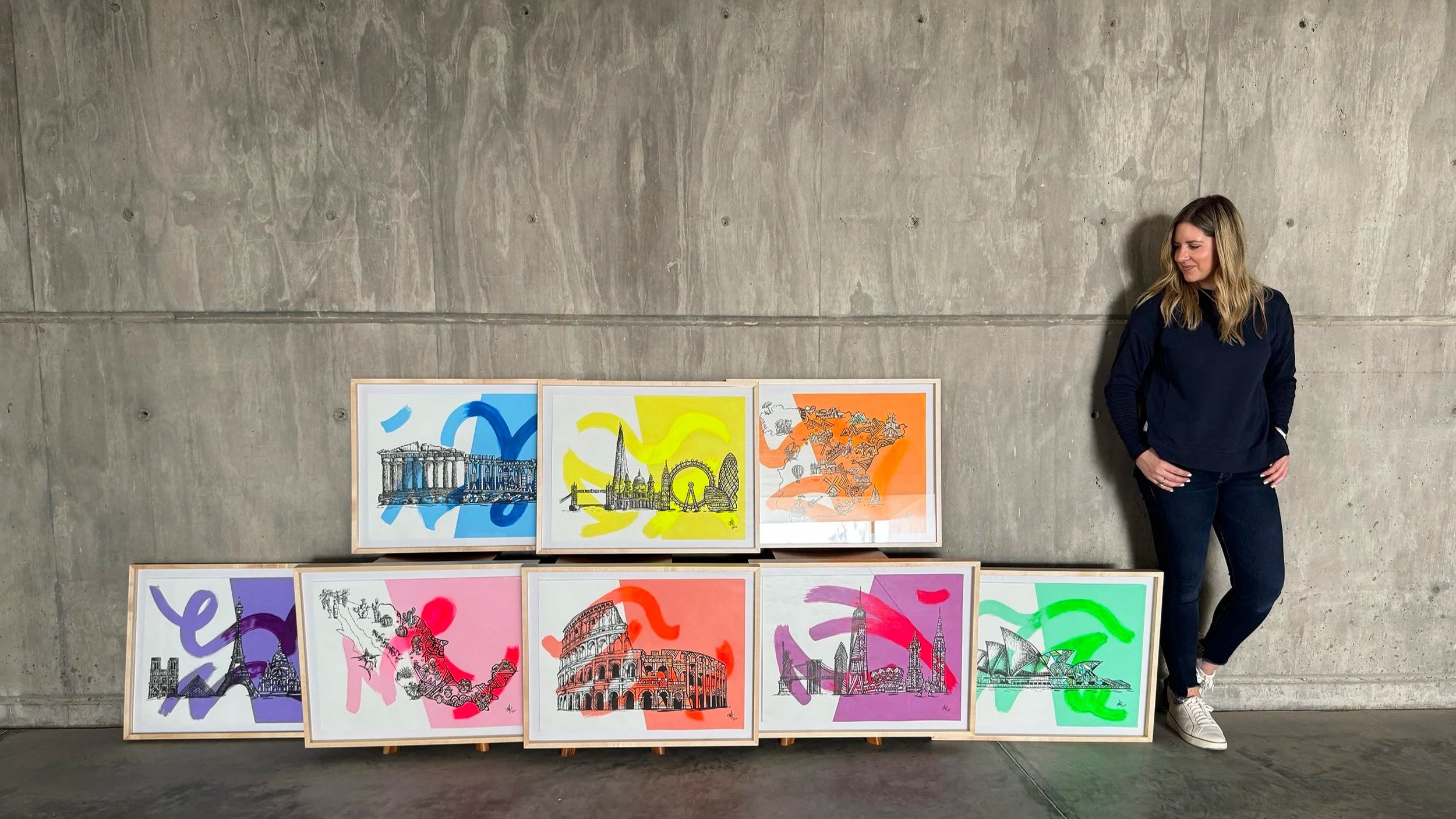 A woman standing with her hands on her hips next to eight colorful framed pictures of famous landmarks, displayed on the floor against a concrete wall. The pictures feature artistic illustrations with vibrant backgrounds in blue, yellow, orange, purple, pink, and green.