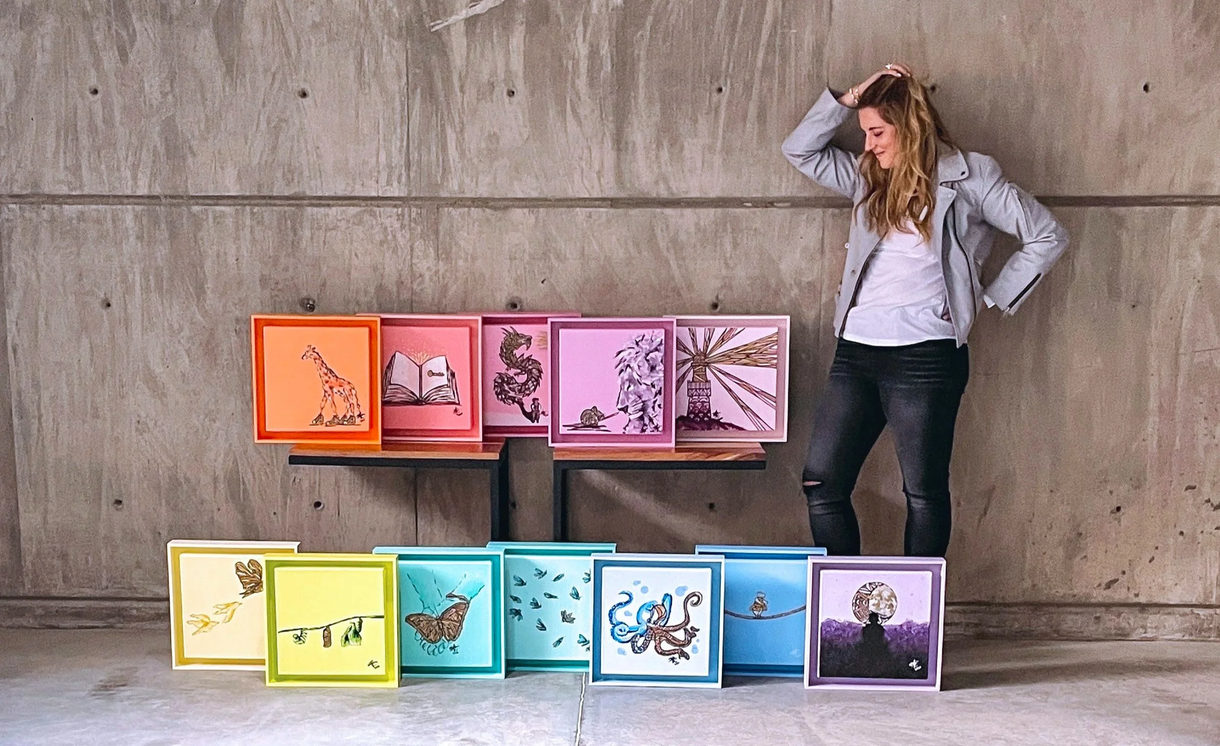 A woman with long wavy hair, wearing a gray jacket and black ripped jeans, stands next to a display of colorful framed artwork against a concrete wall. The artwork features various drawings, including animals, books, and abstract designs in vibrant colors.