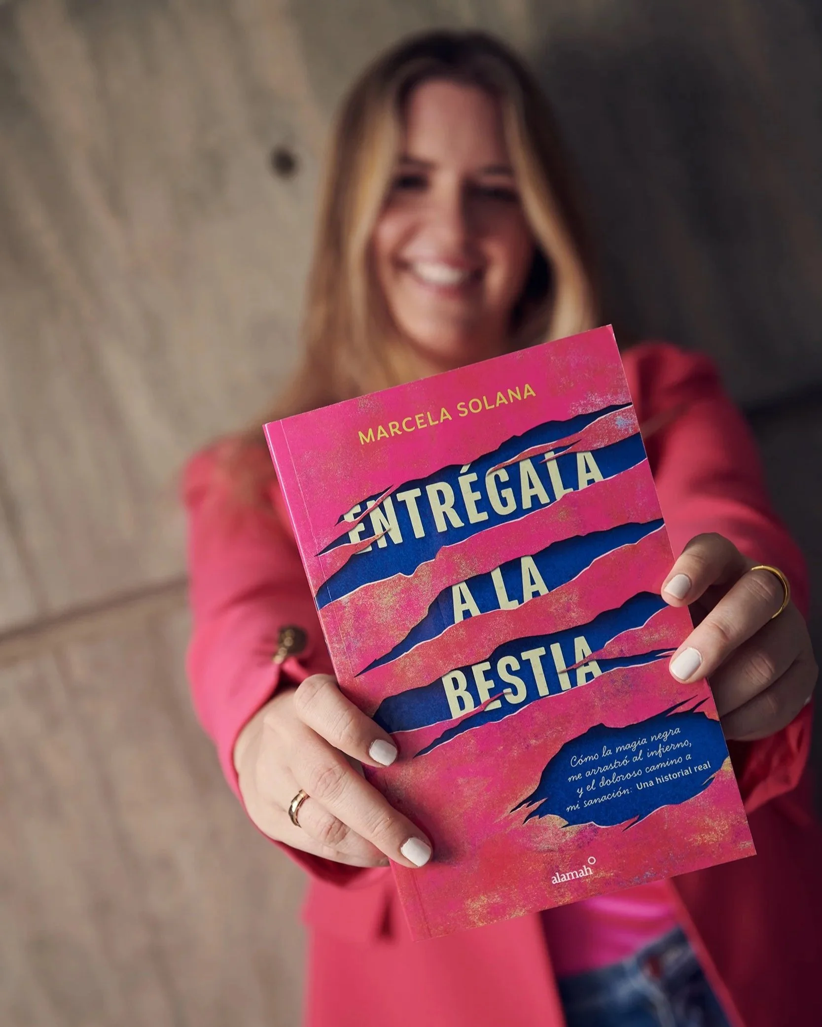 A woman with long hair and a pink jacket smiling and holding a pink book titled 'Entregala a la Bestia'.