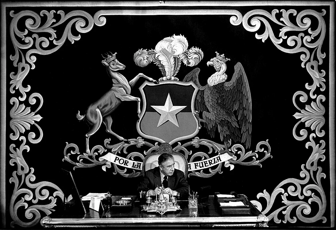 President Augusto Pinochet in his office at the presidential palace. The tapestry behind him reads "BY FORCE", Santiago de Chile, 1975.