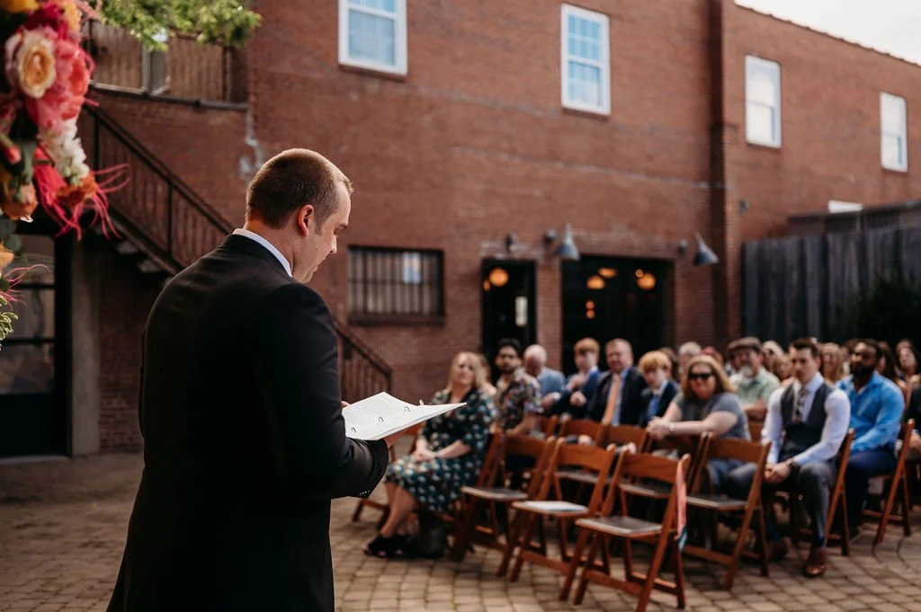  Man reading to a group of people seated in an outdoor courtyard 