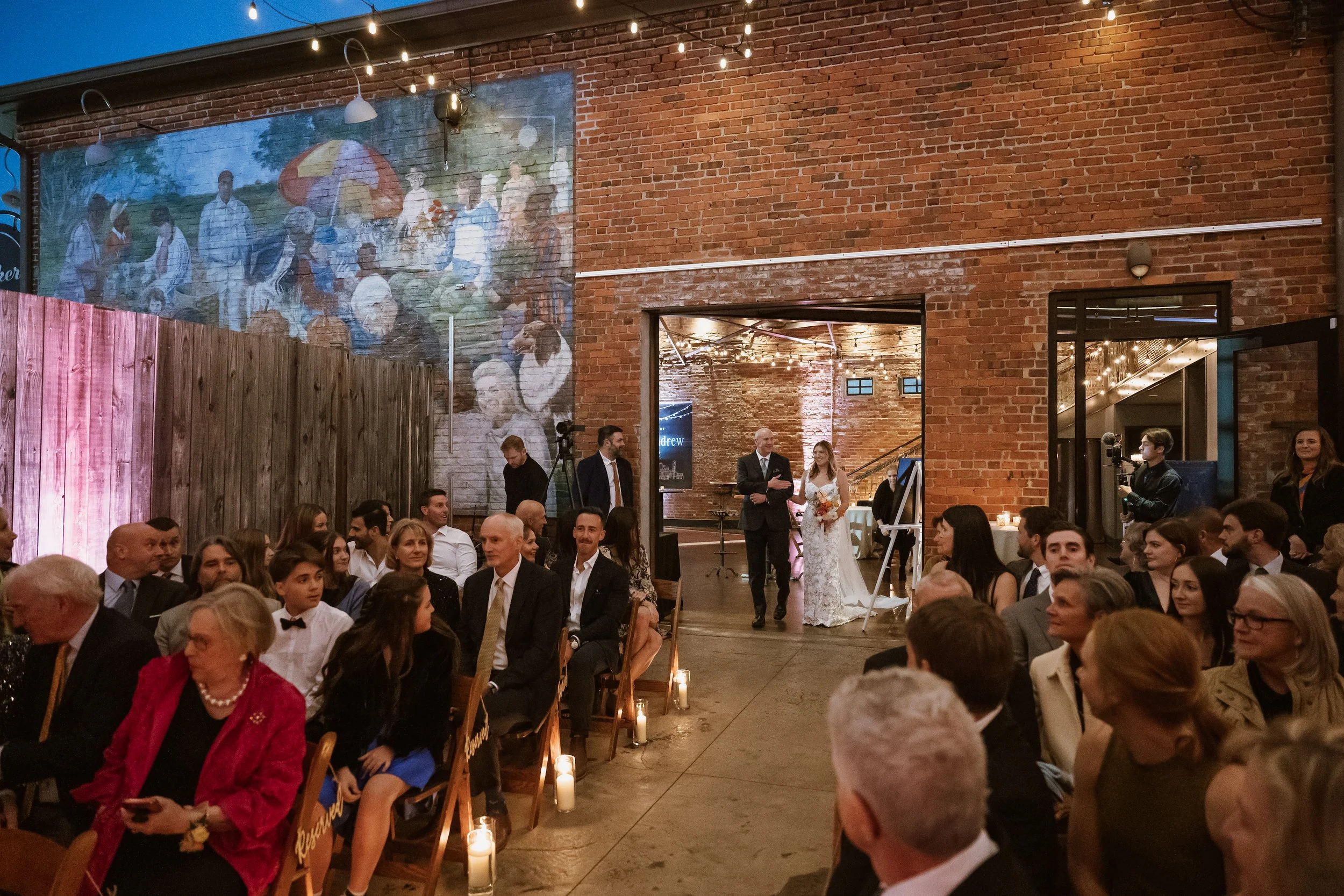 Bride entering outdoor courtyard with seated wedding guests and candlelit aisle