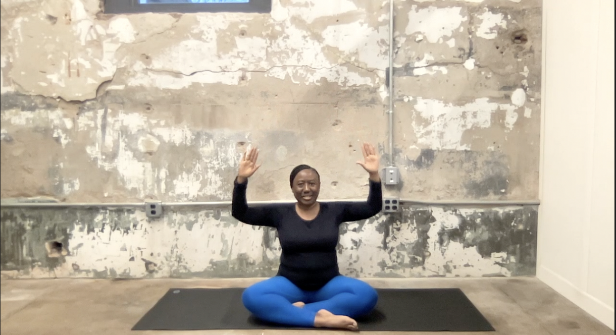Ivory Howard sits on yoga mat with crossed legs and arms in the air.
