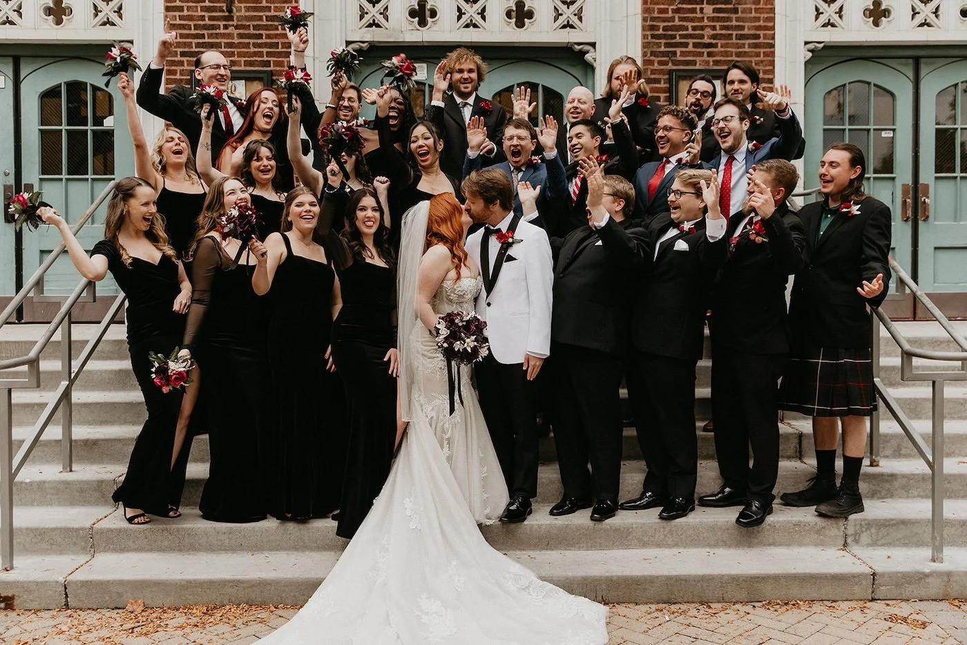 A groom in a white tuxedo and a bride in a mermaid gown kiss in front of their large, cheering bridal party who wear black gowns and black tuxes
