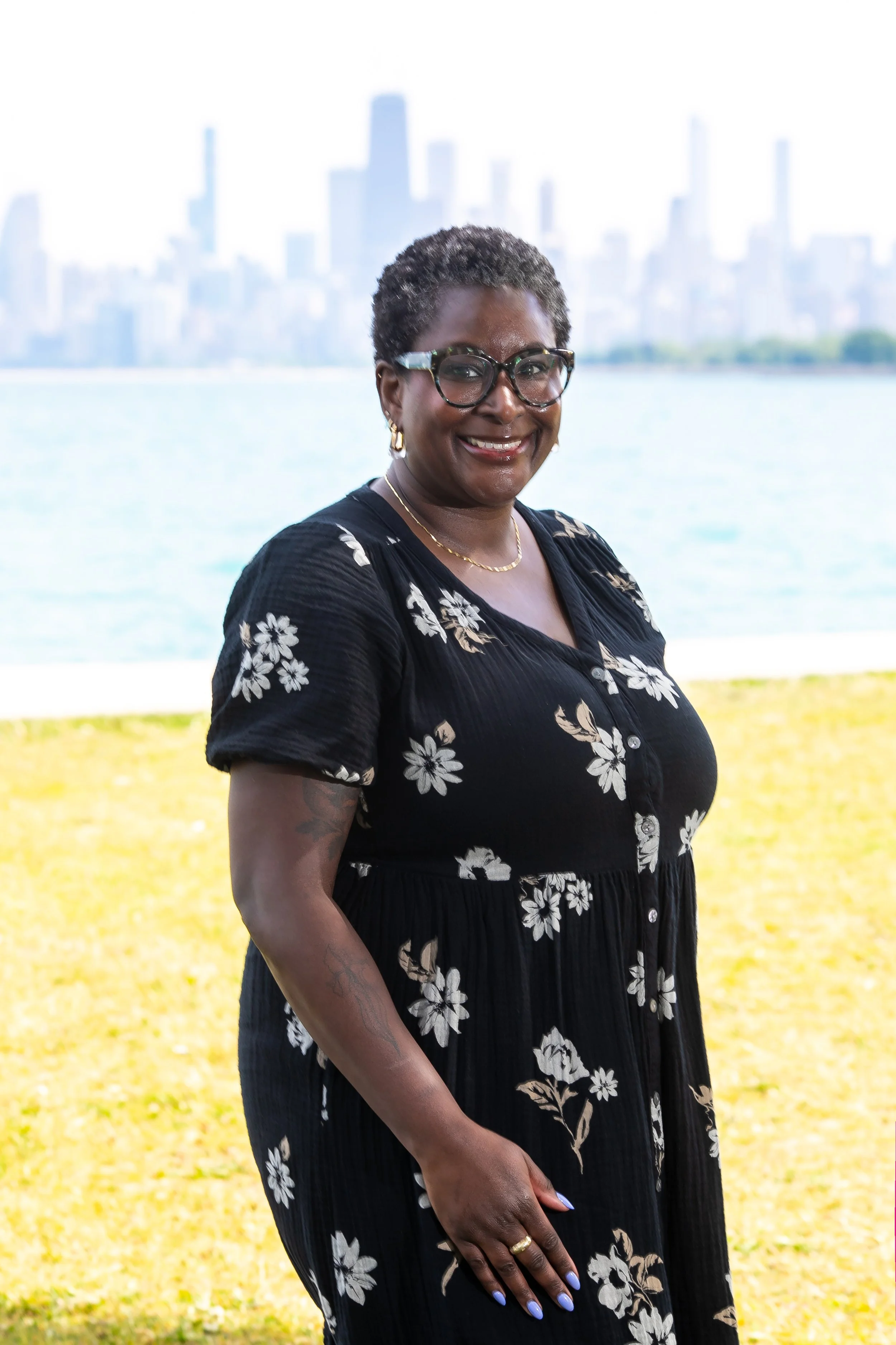 a Black woman in a black flowered dress stands on the grass smiling in front of Lake Michigan and the Chicago skyline