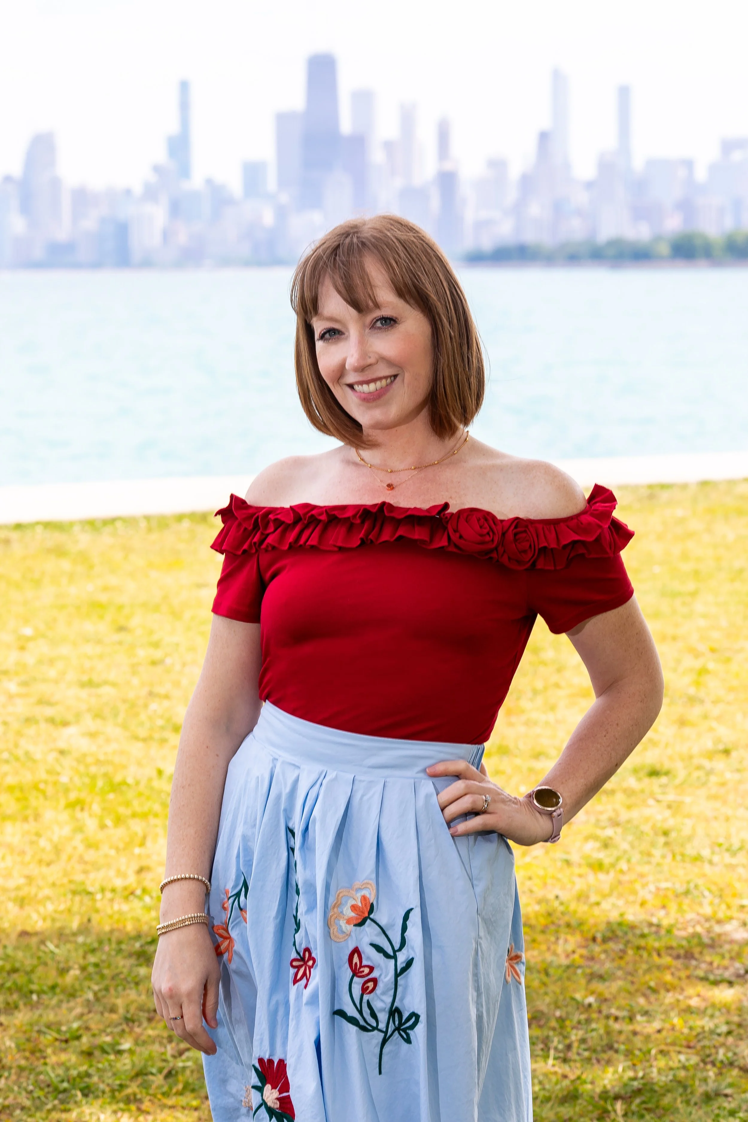 a redheaded white woman wedding planner and coordinator stands in a red top and blue skirt in front of the chicago skyline