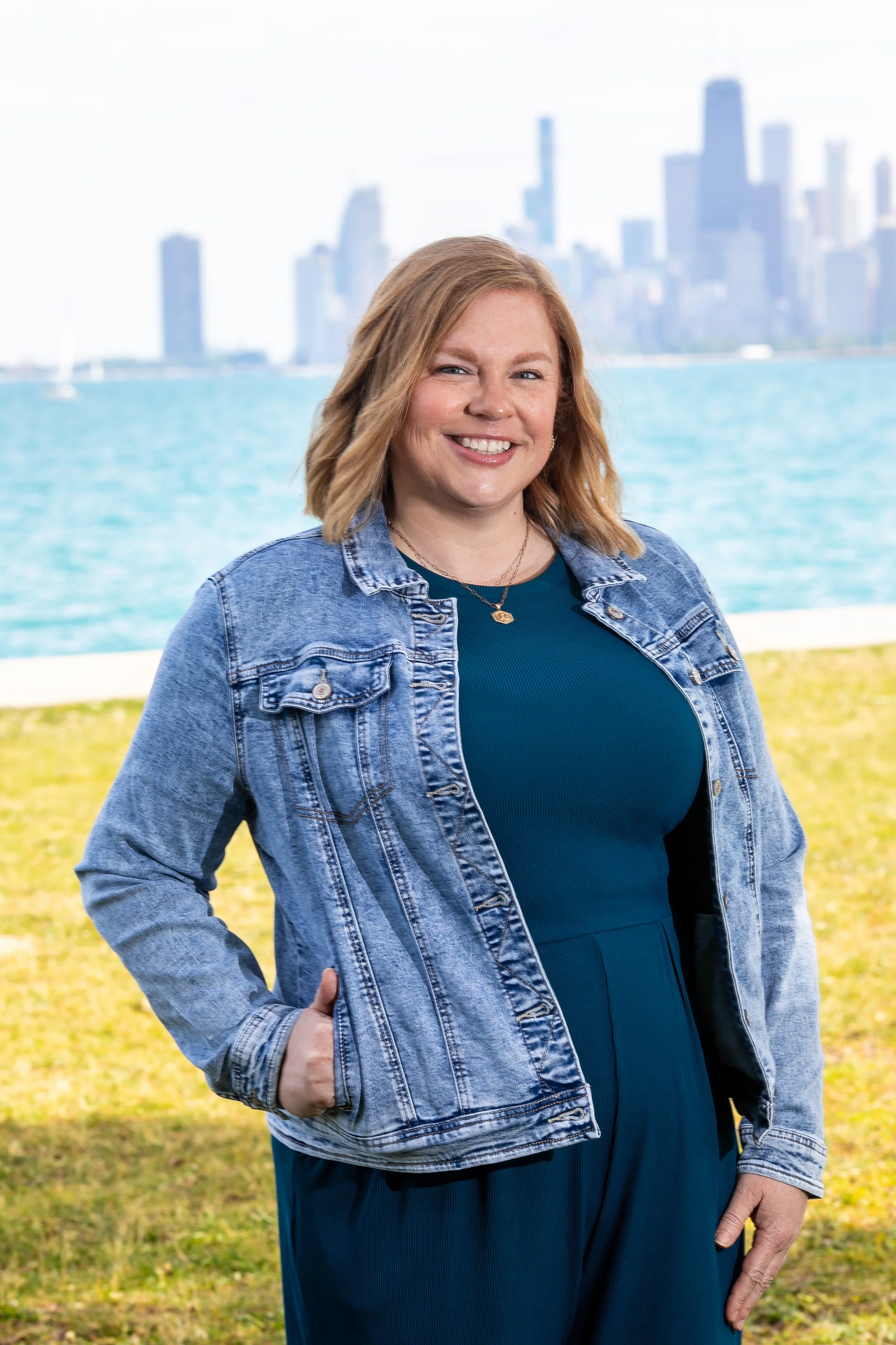 a redheaded white woman in a jean jacket and blue jumpsuit stands on the grass smiling in front of Lake Michigan and the Chicago skyline