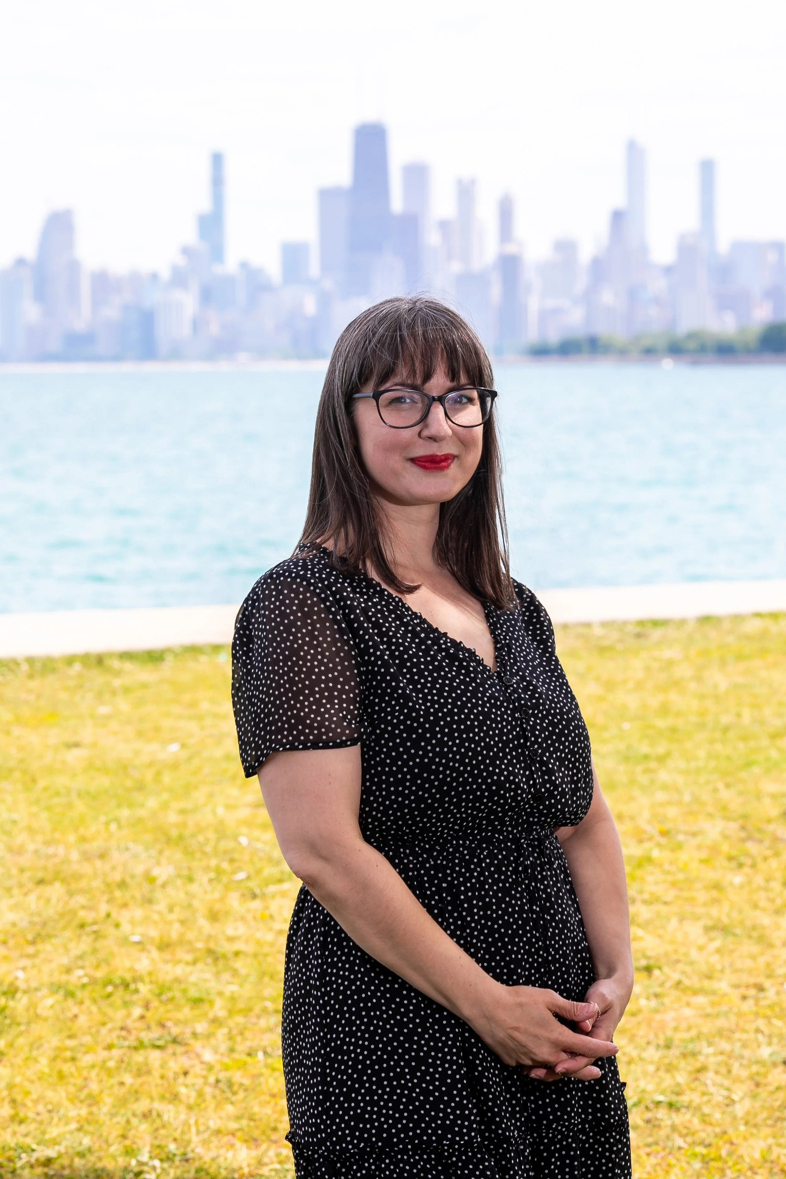 a white brunette assistant wedding coordinator in glasses and red lipstick stands in front of the chicago skyline in a black dress