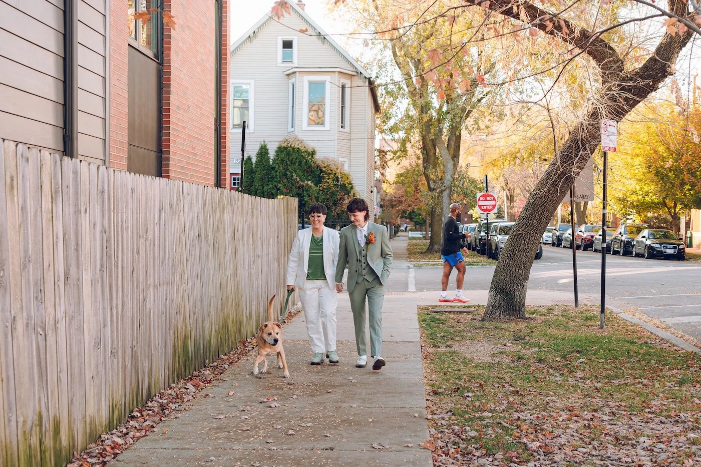 two brides in a white suit and a green suit walk down a street in chicago walking their dog