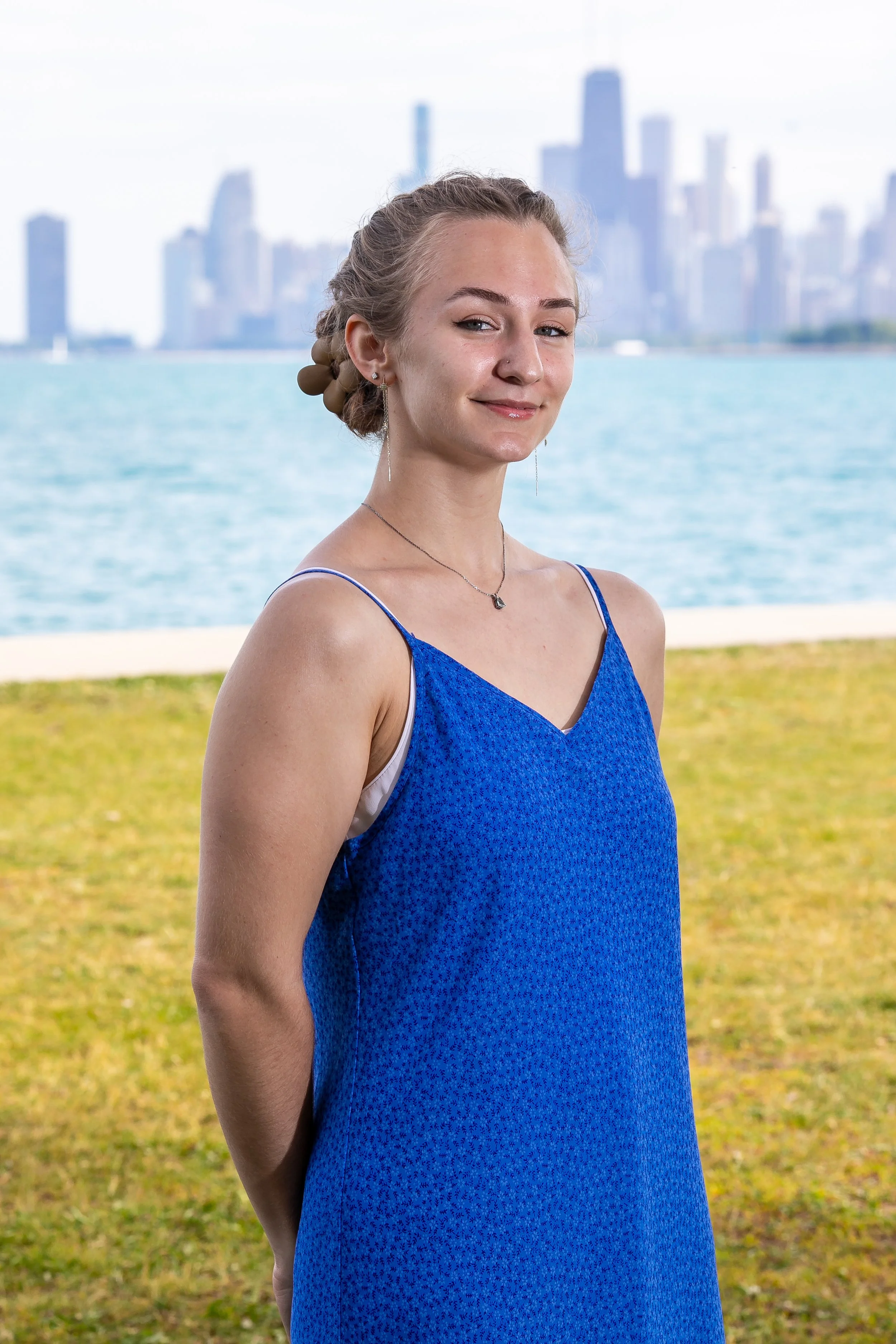 a blonde white person in a strappy blue dressstands on the grass smiling in front of Lake Michigan and the Chicago skyline