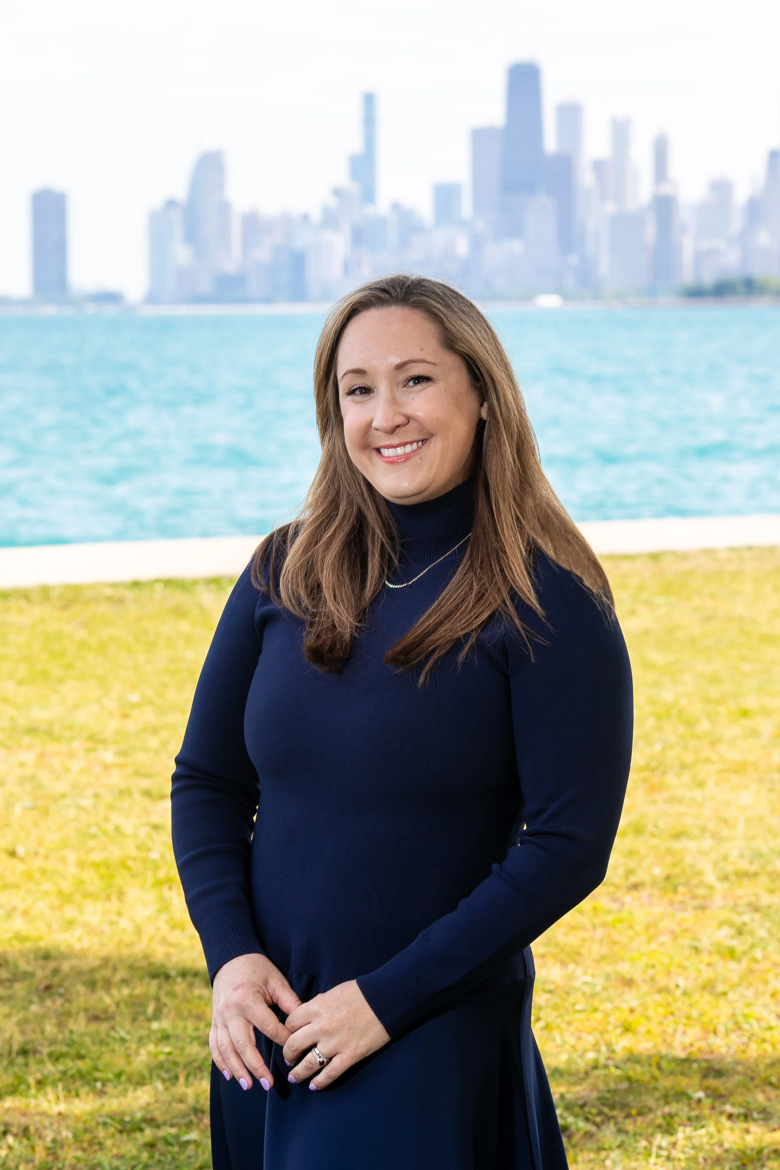 a brunette white woman in a navy dress stands on the grass smiling in front of Lake Michigan and the Chicago skyline