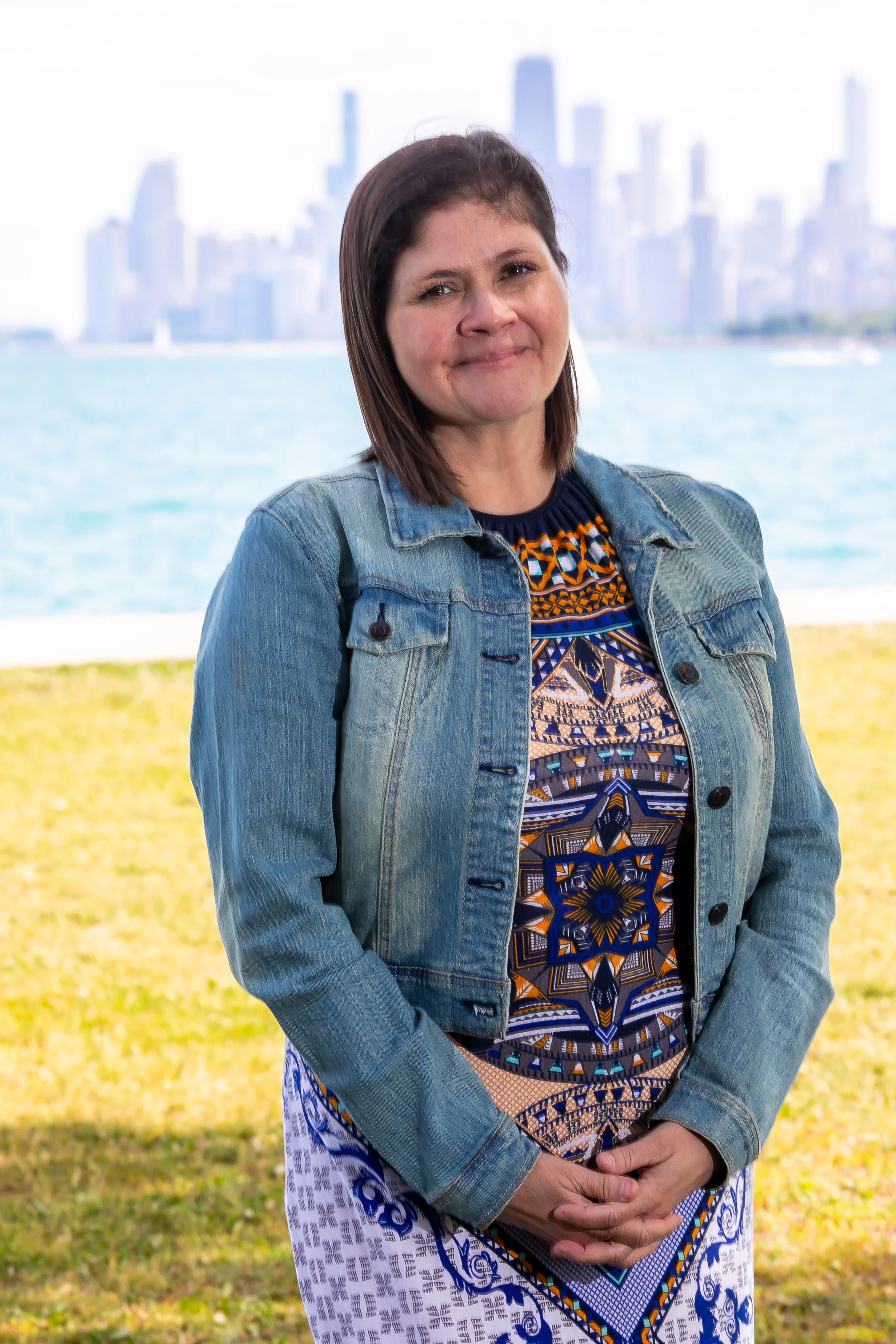 a white brunette woman in a jean jacket stands on the grass smiling in front of Lake Michigan and the Chicago skyline