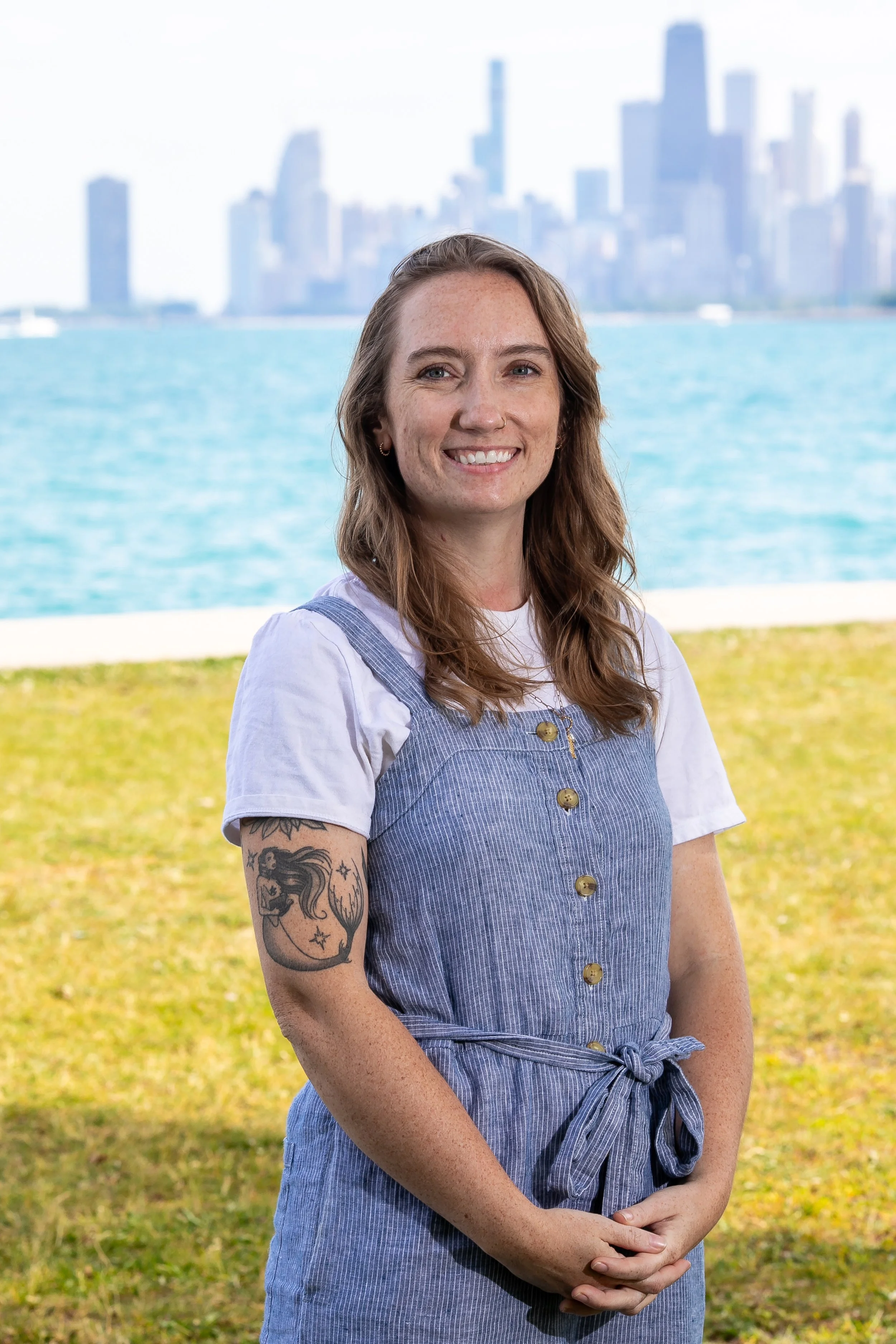 a brunette white woman wearing striped overallsstands on the grass smiling in front of Lake Michigan and the Chicago skyline
