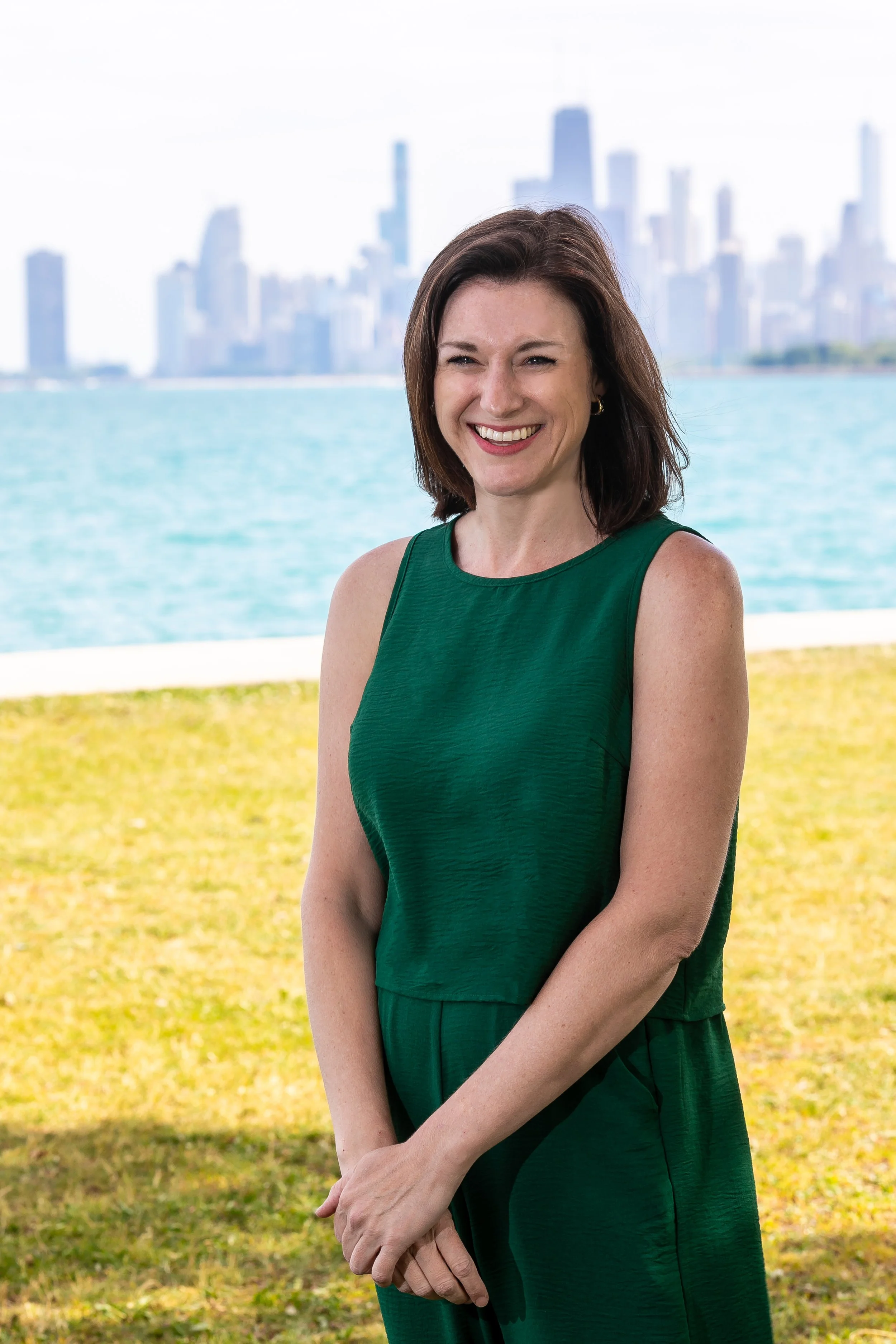 brunette white woman wedding planner wearing green stands on the grass in front of the chicago skyline on Lake Michigan