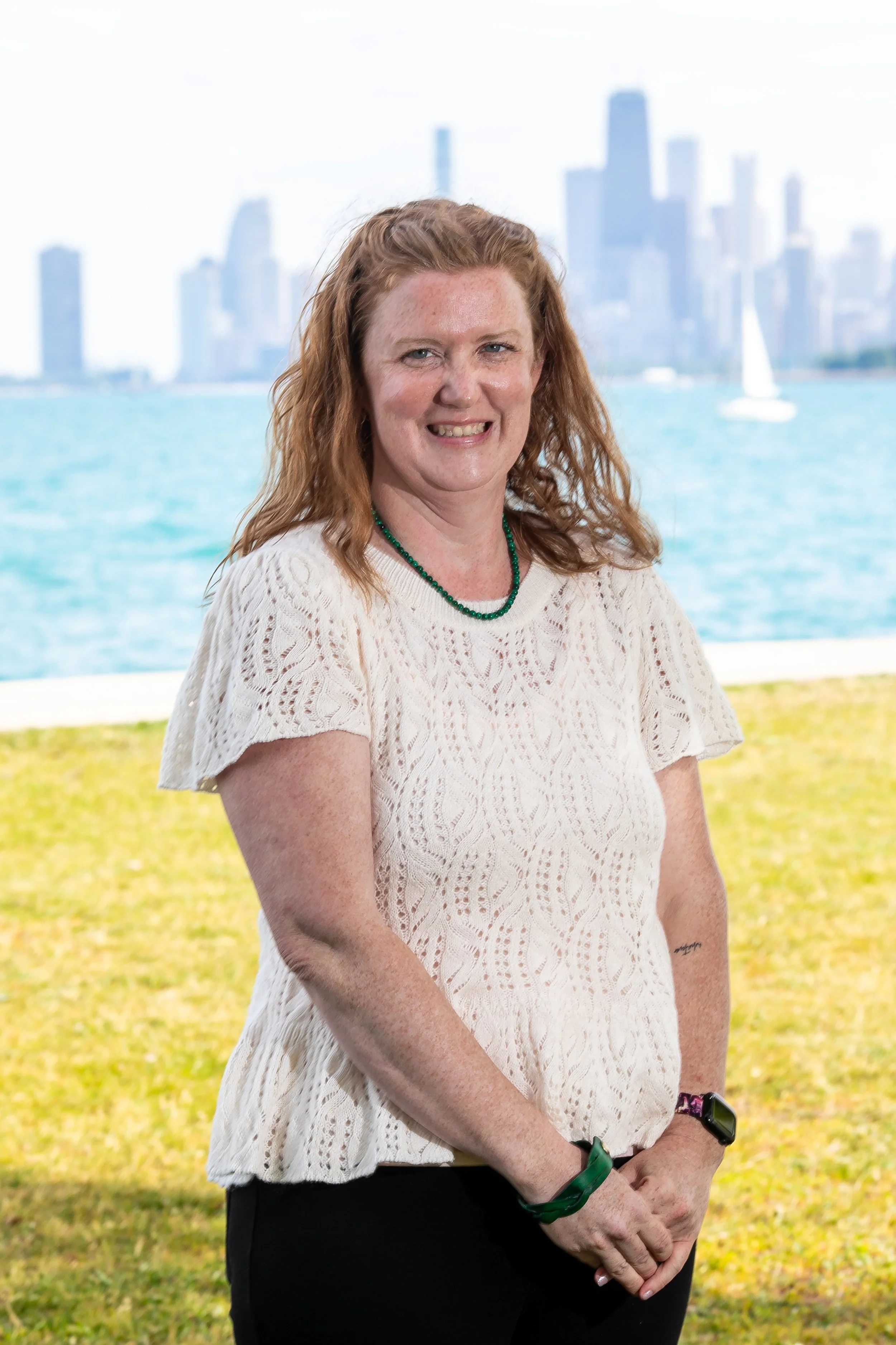 a redheaded white woman wearing a white top stands on the grass smiling in front of Lake Michigan and the Chicago skyline